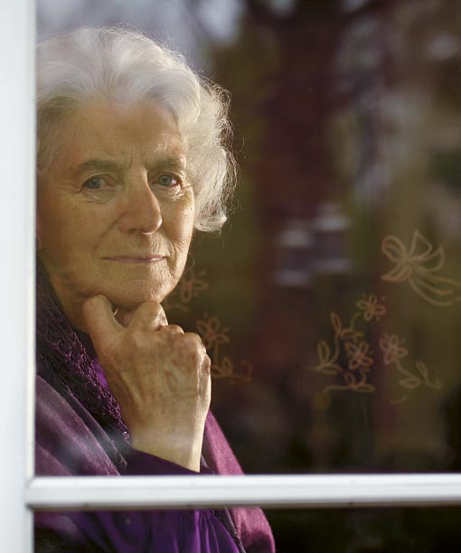 Grethe Engelbrecht med hvidt hår og lilla tøj, der ser tankefuldt ud gennem et vindue med blomsterdekorationer.