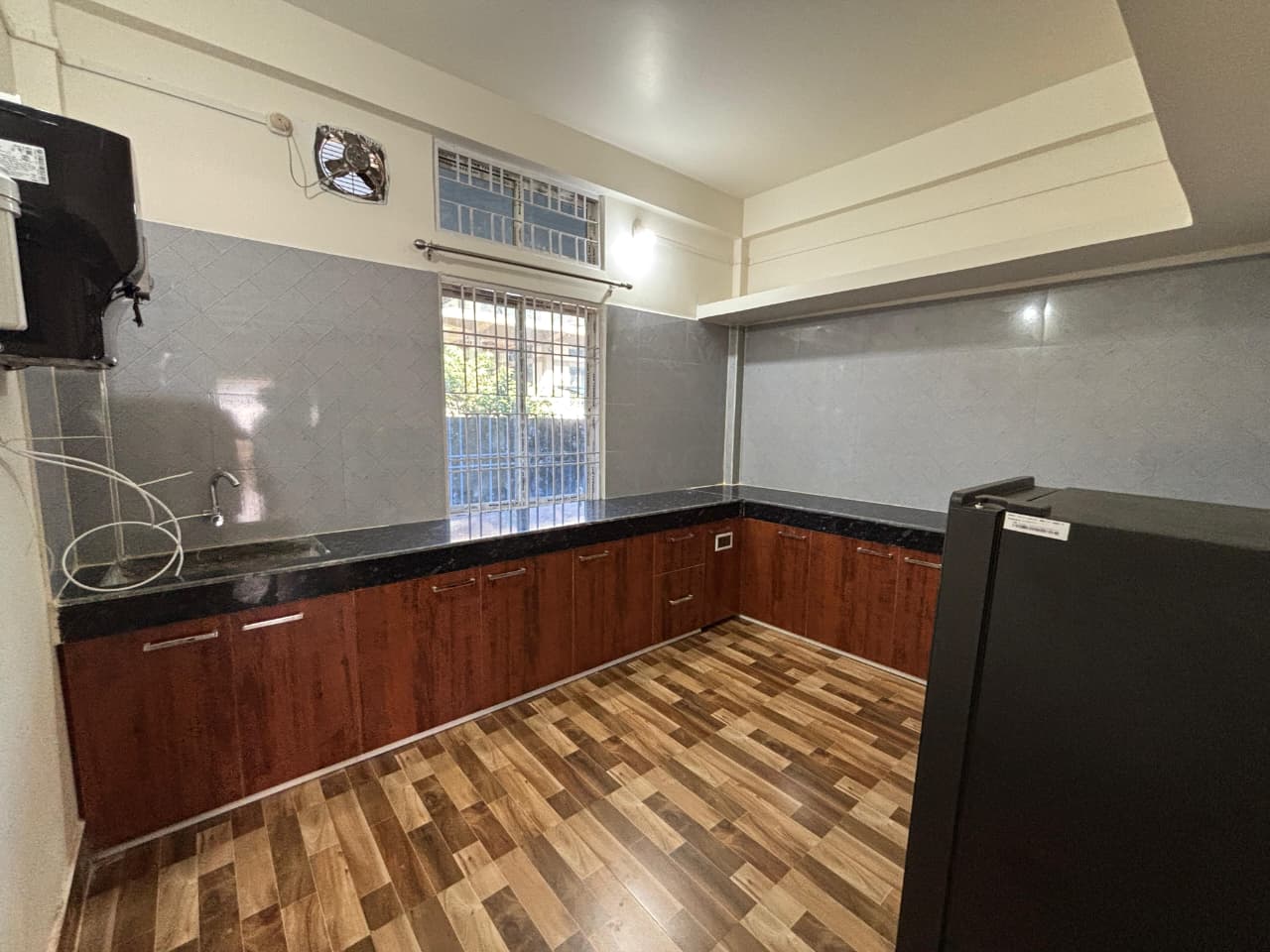 Empty kitchen with L-shaped black countertop, wooden cabinets, a window with metal bars, and a black refrigerator on the right.