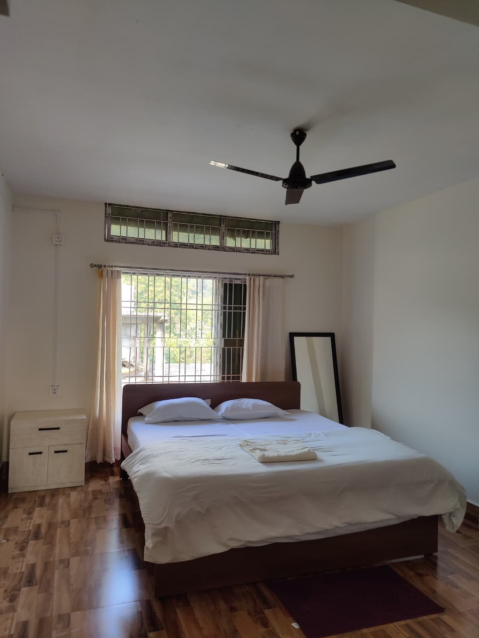 Minimalist bedroom with a double bed featuring white bedding, two pillows, a folded towel on top, a wooden nightstand, a standing mirror, and a ceiling fan.