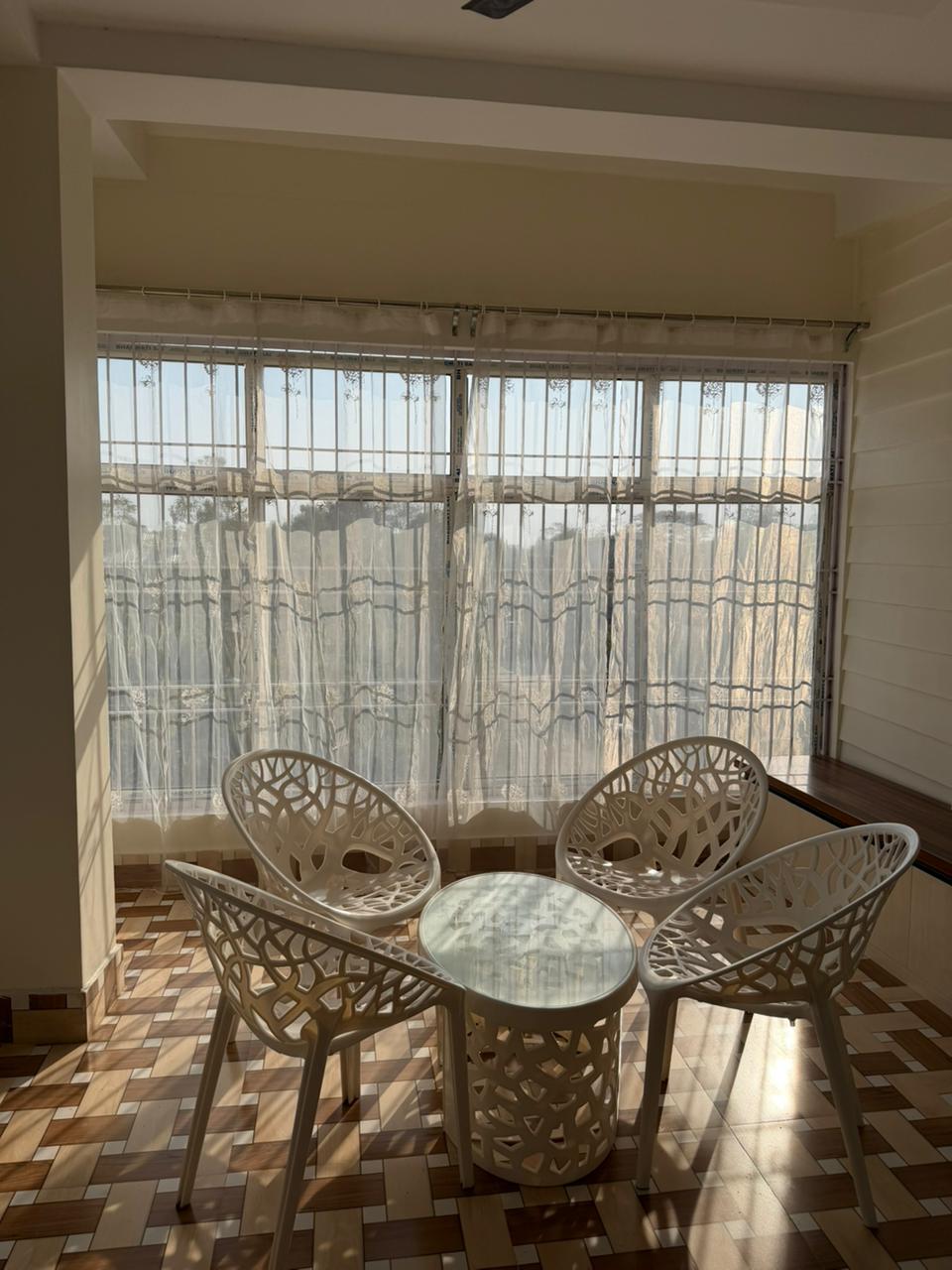 Sunlit corner with four white decorative chairs arranged around a small round glass-top table by a window with sheer patterned curtains.