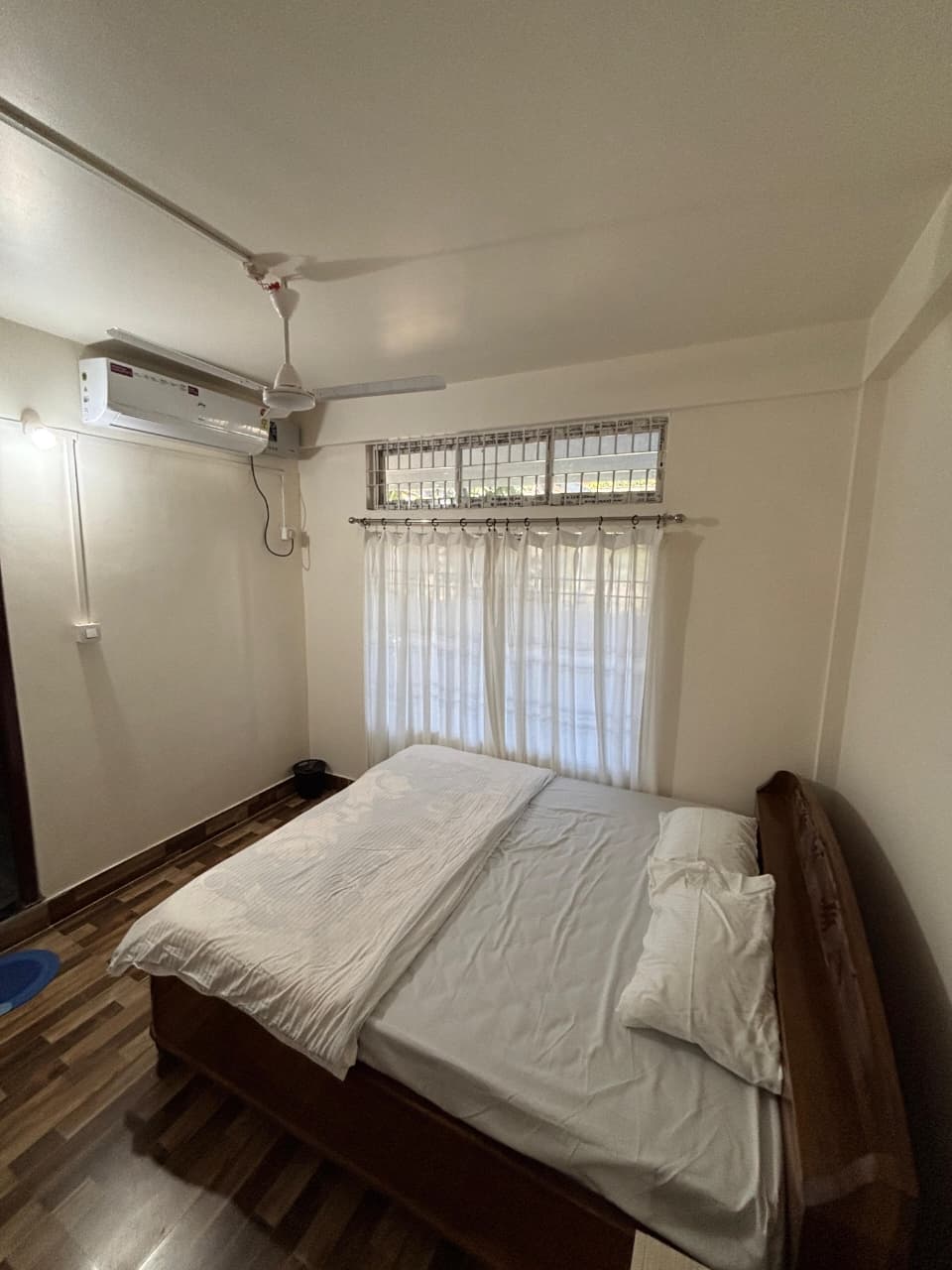 Minimalist bedroom with a wooden bed, white bedding, sheer curtains, ceiling fan, and wall-mounted air conditioner.