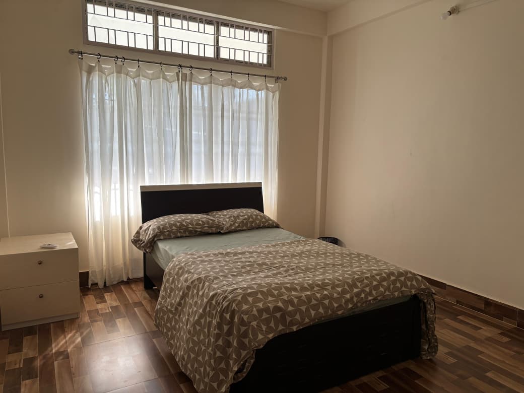 Minimal bedroom with a bed, patterned beige and white bedding, sheer white curtains, and a wooden nightstand on a wood floor.