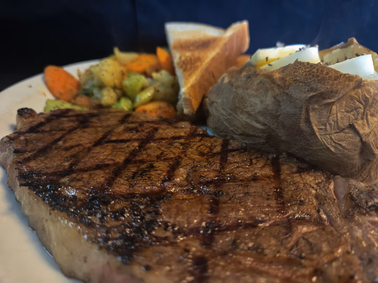 Grilled steak with crosshatch marks, served with a baked potato topped with butter and a side of mixed vegetables.