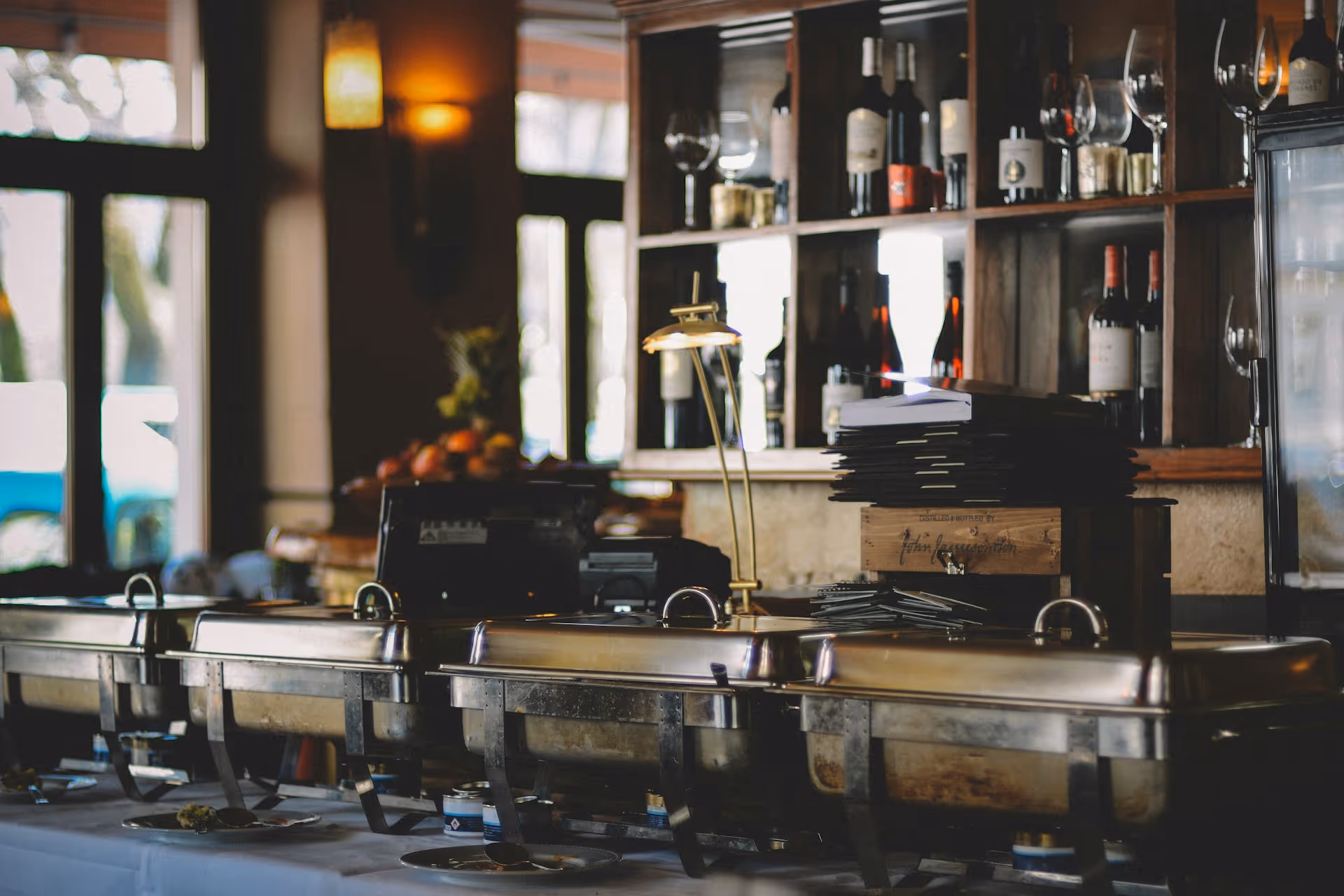 Buffet setup in a restaurant with warm lighting. Silver chafing dishes line a table, and wine bottles fill shelves.
