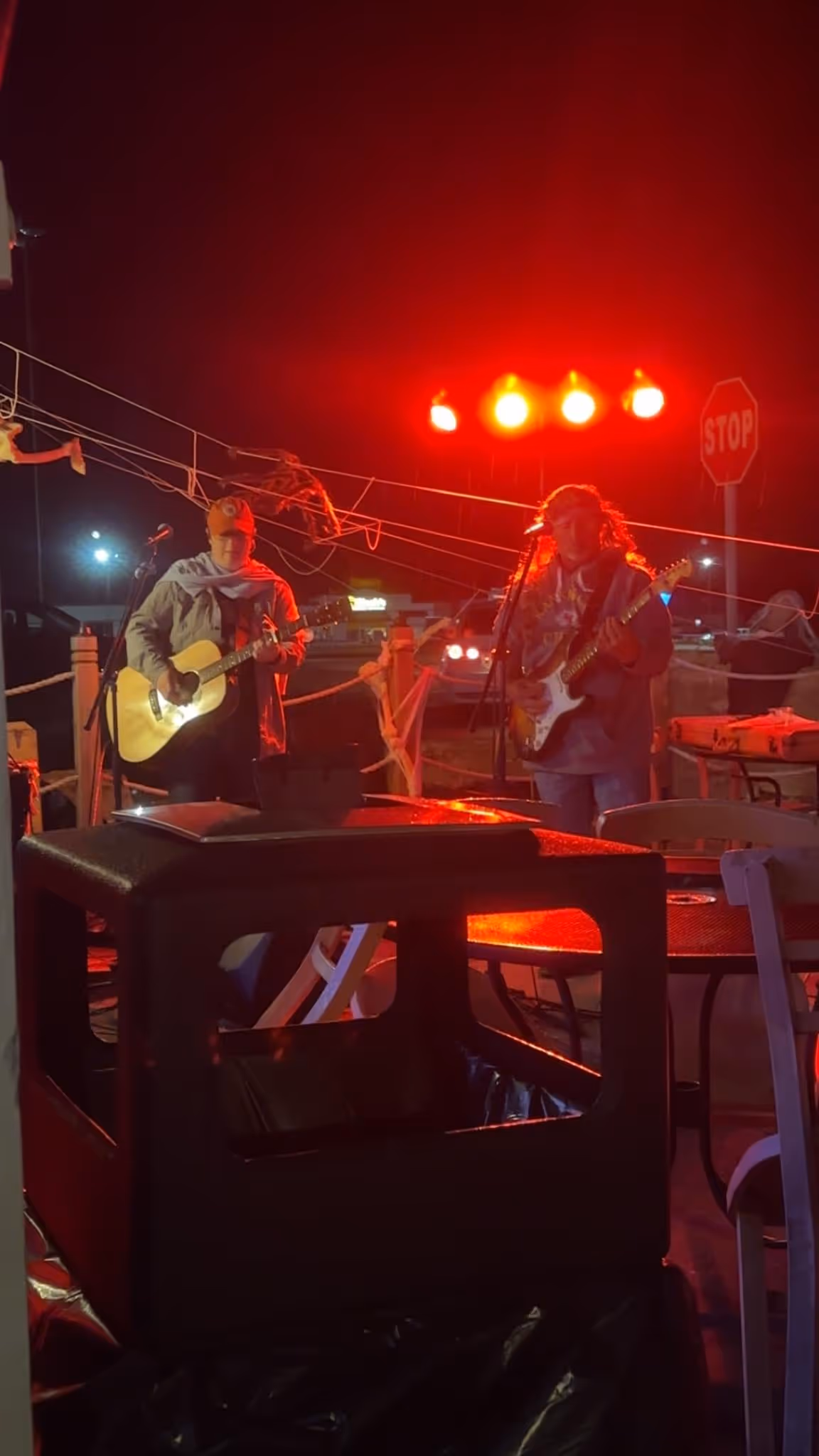 Two musicians perform outdoors at night under red stage lights. One plays an acoustic guitar, the other an electric. A stop sign is visible nearby.