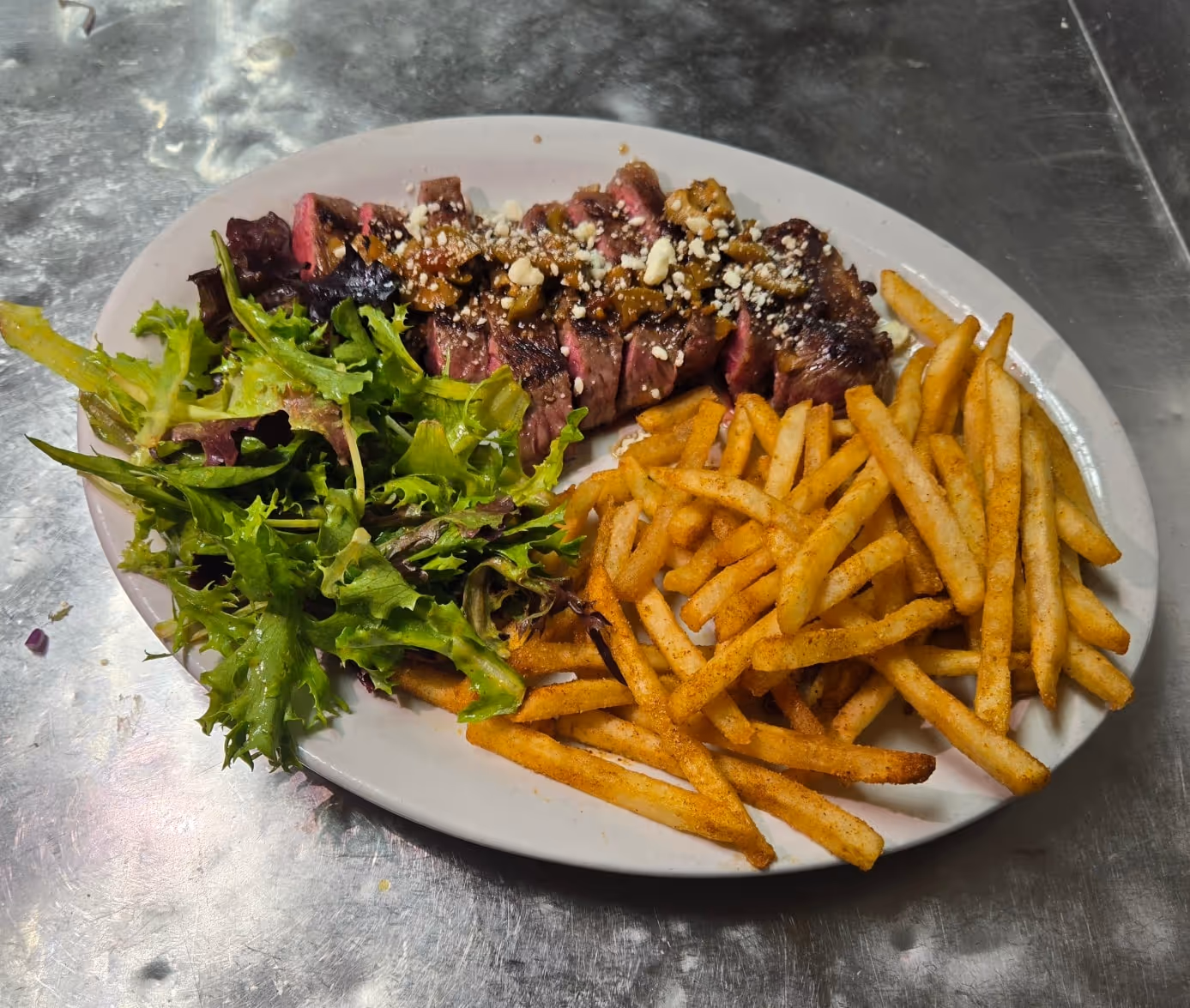 A plate of sliced steak with crumbled cheese and sauce, served with crispy fries and fresh mixed greens. It conveys a hearty, appetizing meal.
