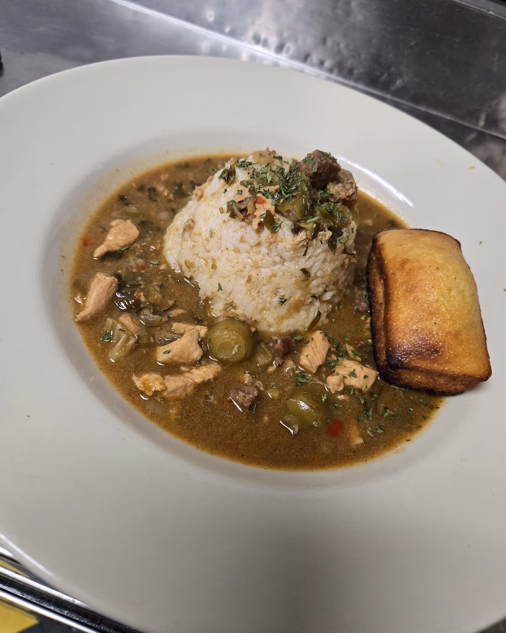 A dish of gumbo featuring a mound of white rice in the center, surrounded by a savory broth with vegetables and meat chunks, garnished with herbs. A piece of cornbread sits on the side, creating a warm, hearty meal ambiance.