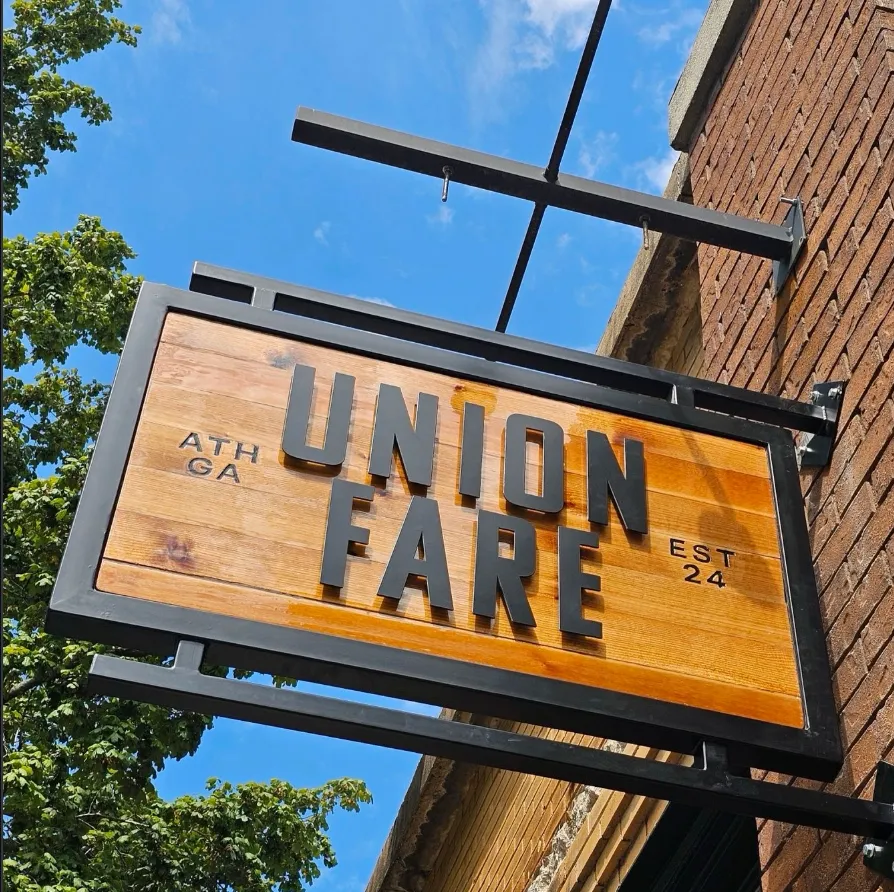 Wooden sign for Union Fare restaurant mounted on brick wall with blue sky and green trees background.