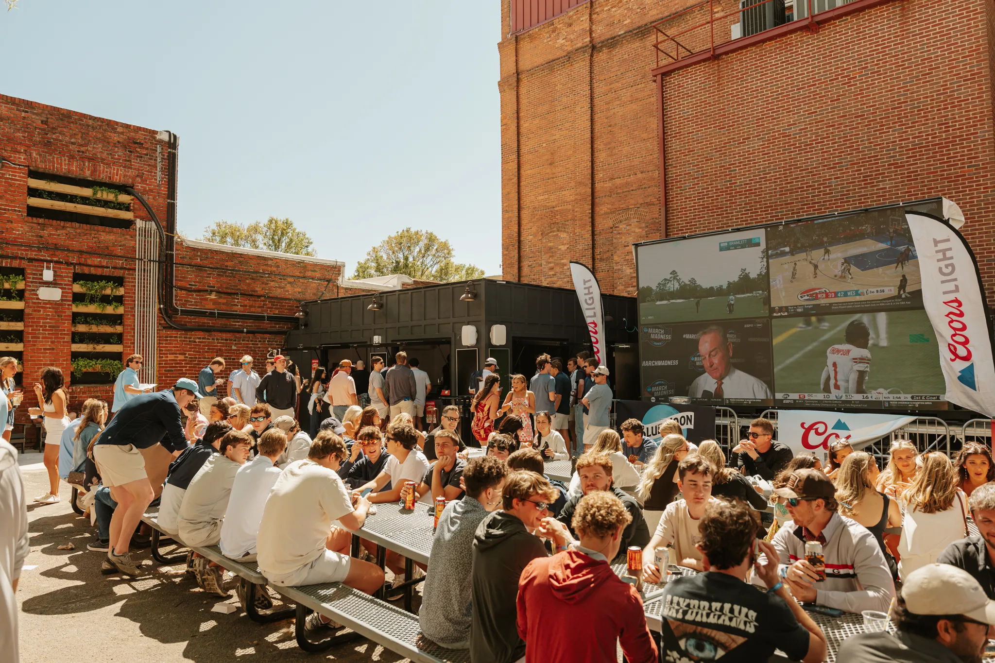 Outdoor crowd seated at metal tables watching sports on large screen with Coors Light banners.