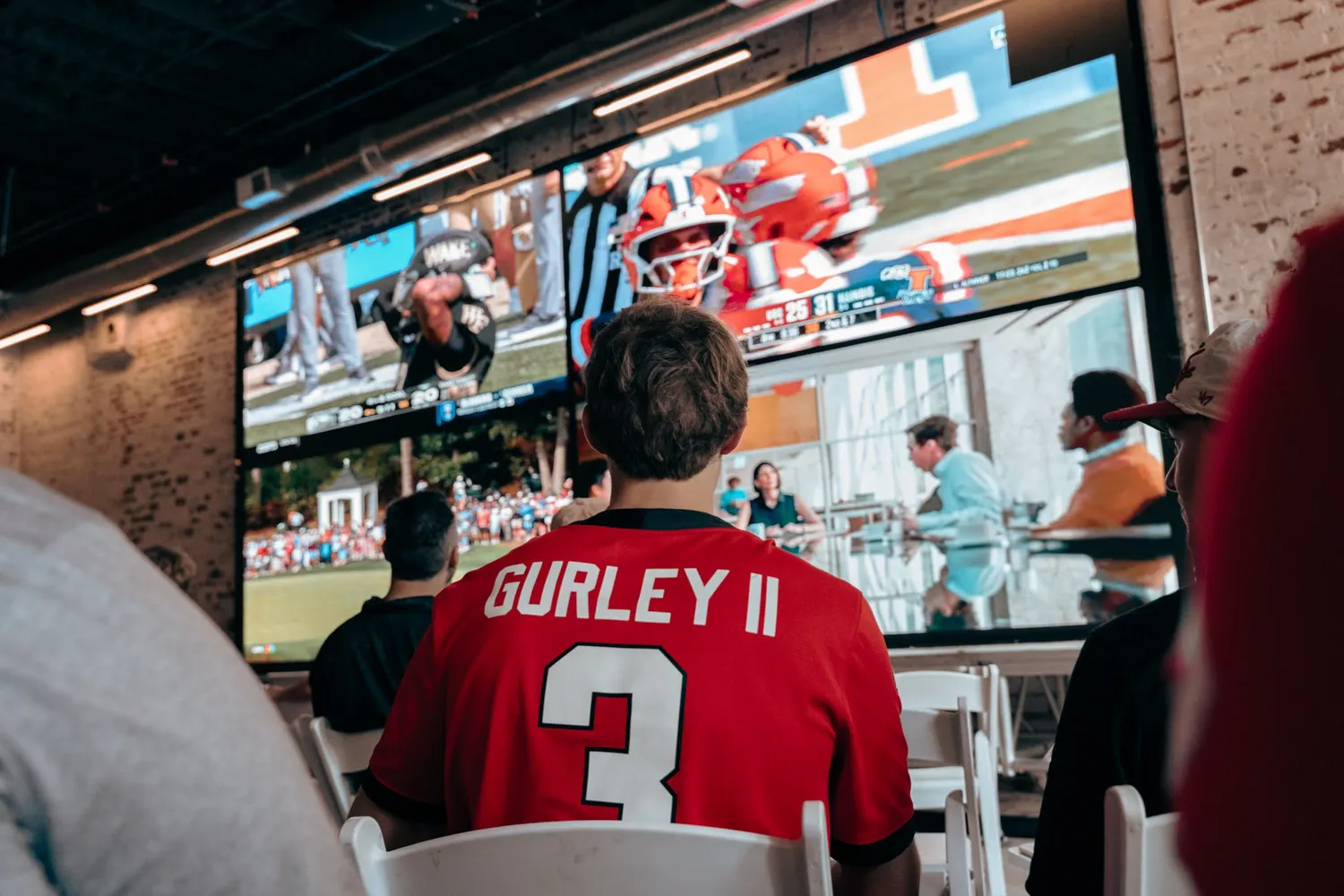 Man wearing red Gurley II jersey watches sports game on large split-screen TV in a room.