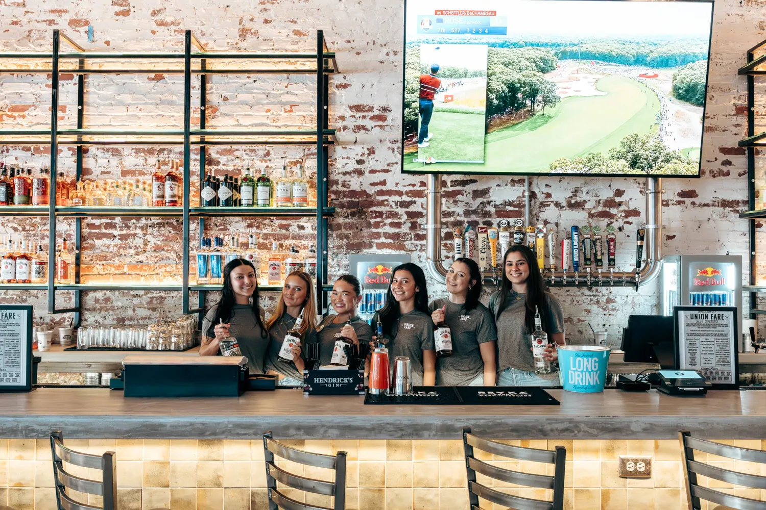Six female bartenders standing behind bar at Union Fare with bottles and golf on TV screen above.