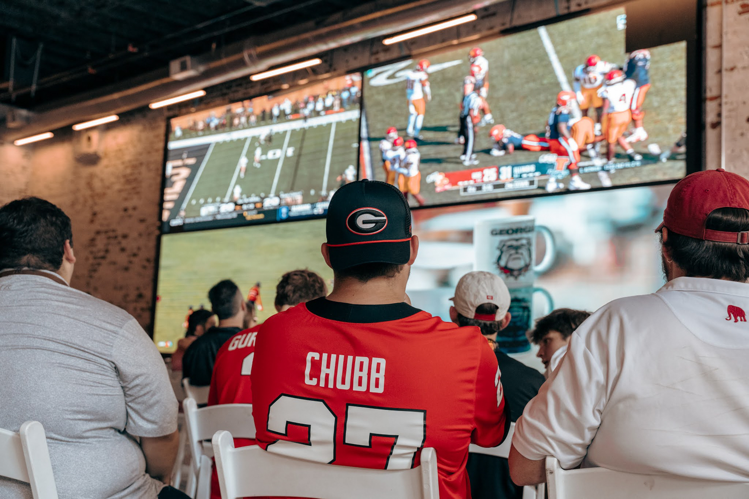 Fans watch a football game on multiple large screens in an indoor bar or lounge.