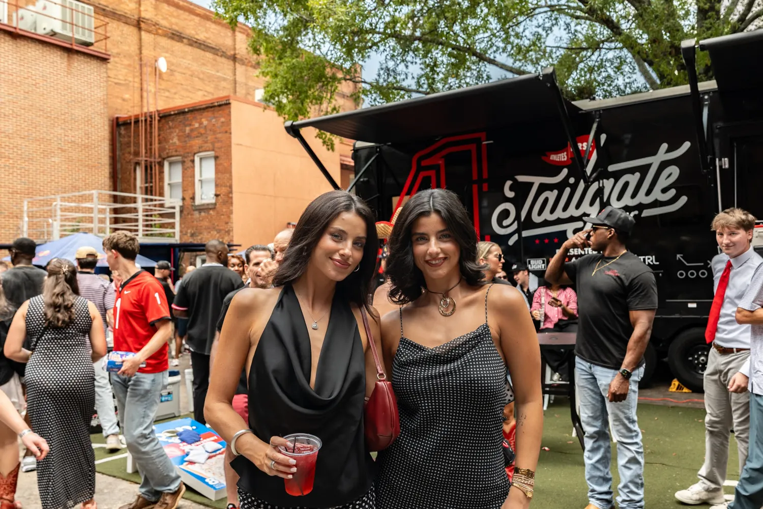 Two smiling women posing at an outdoor tailgate party with a crowd and black tailgate truck in background.