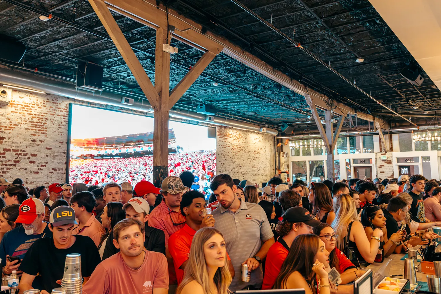 Crowded bar with people watching a large screen showing a sports game in a rustic setting.