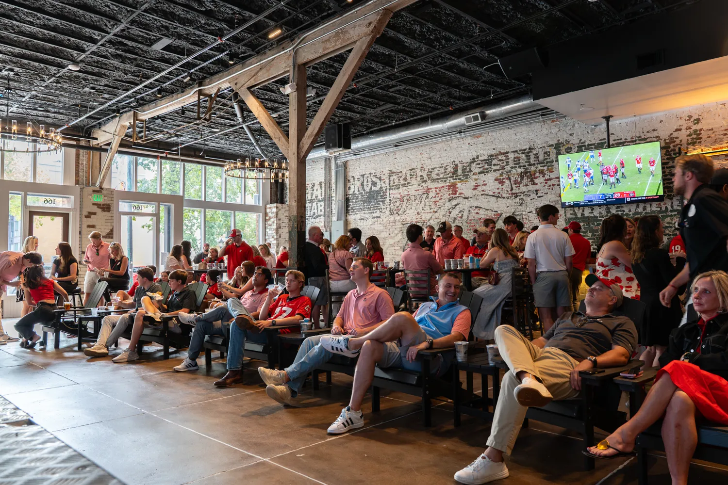 Group of people sitting and standing in a spacious bar watching a football game on TV.
