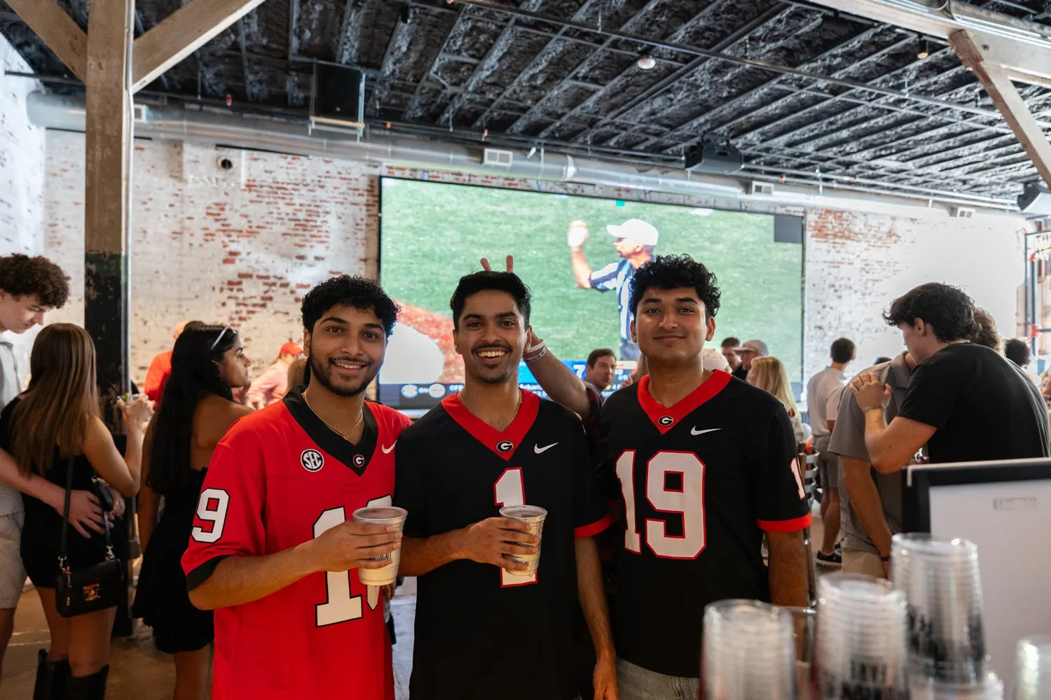 Three young men in Georgia football jerseys holding drinks at a busy indoor sports bar.