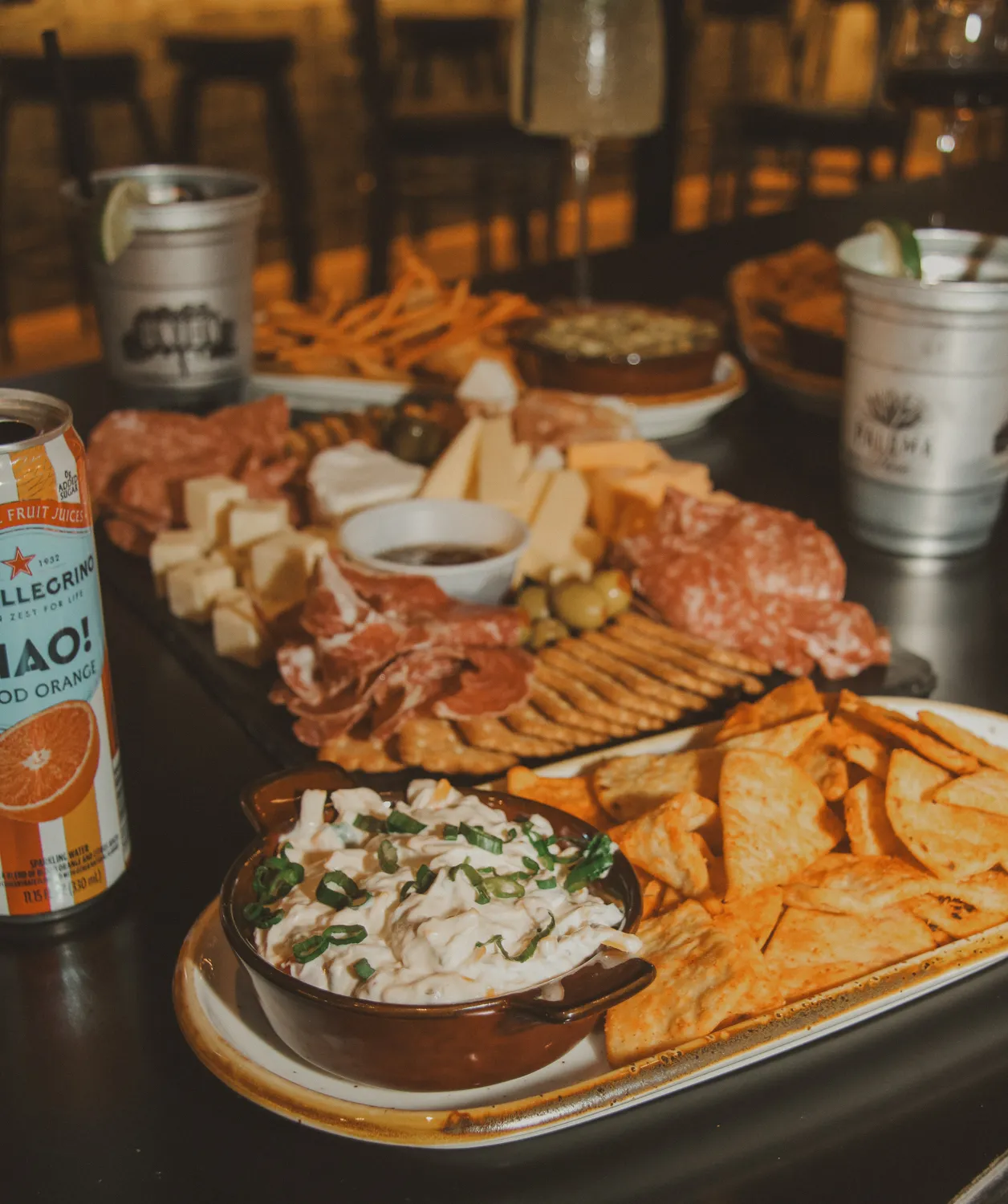 Tray with dip topped with green onions, chips, and a meat and cheese charcuterie board with drinks on table.