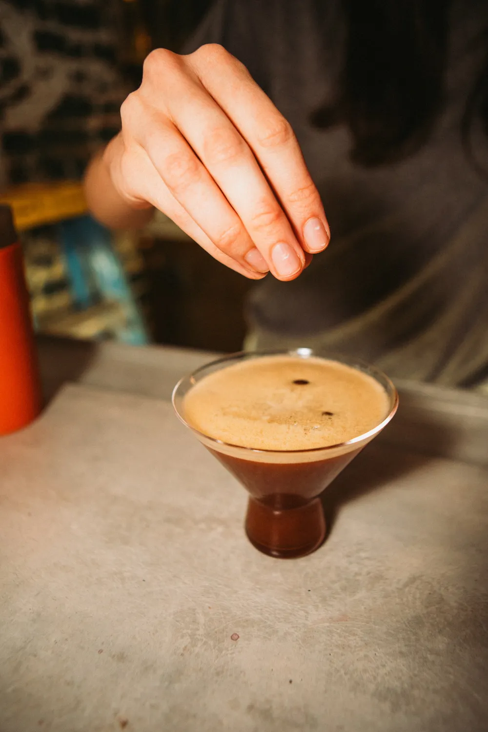 Hand sprinkling garnish over a glass of espresso martini on a table.