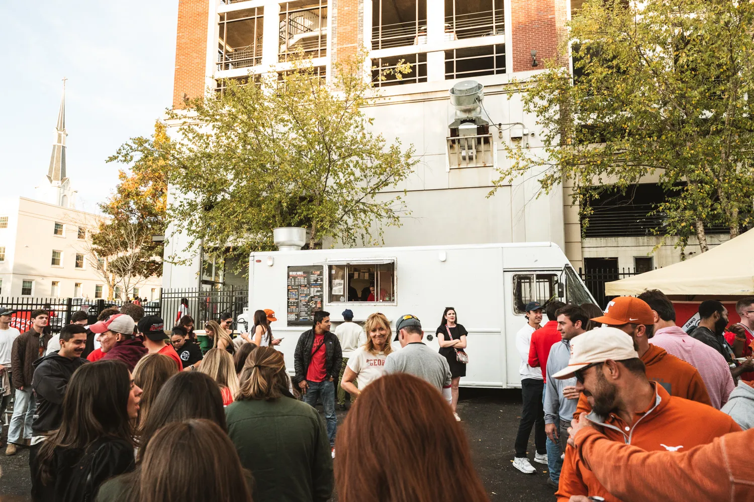 Crowd of people gathered around a white food truck outdoors near a brick building and trees.
