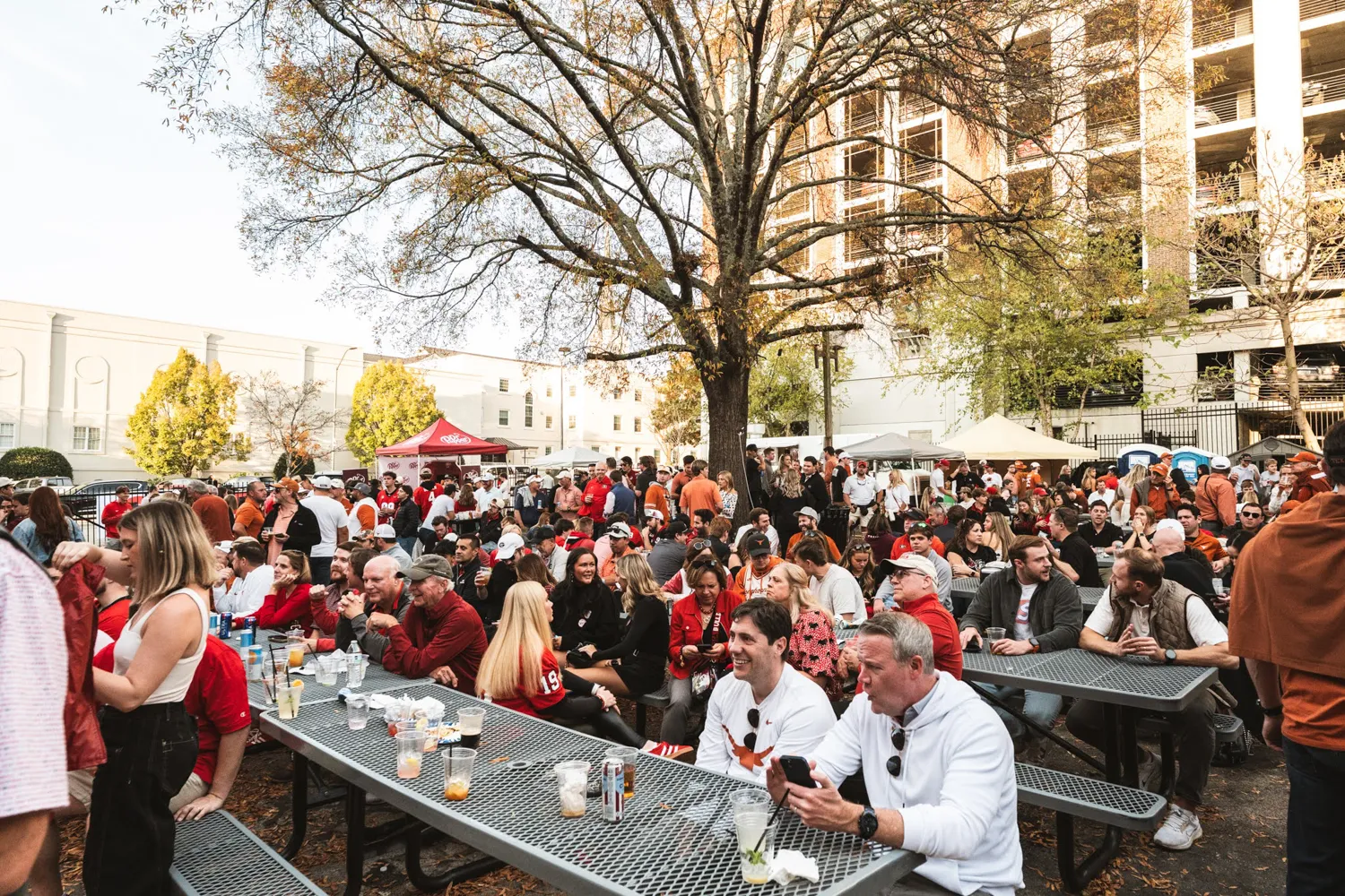 Crowd of people socializing and sitting at picnic tables under a tree in an outdoor event.