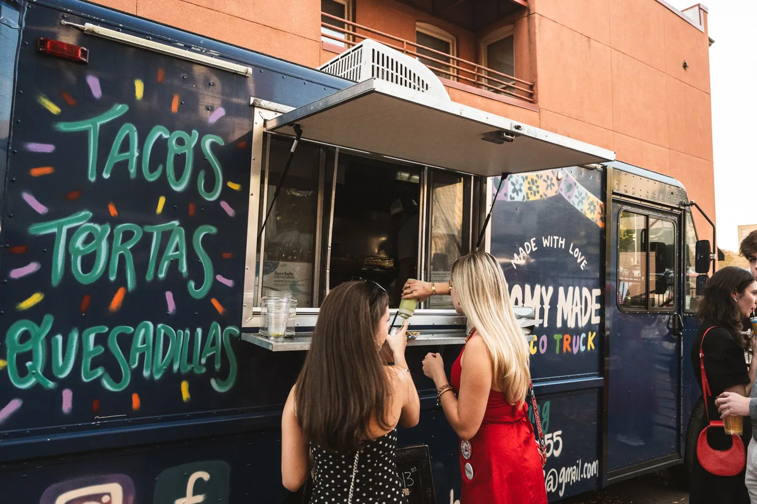 Two women order food at a taco truck with colorful menu words: tacos, tortas, quesadillas.