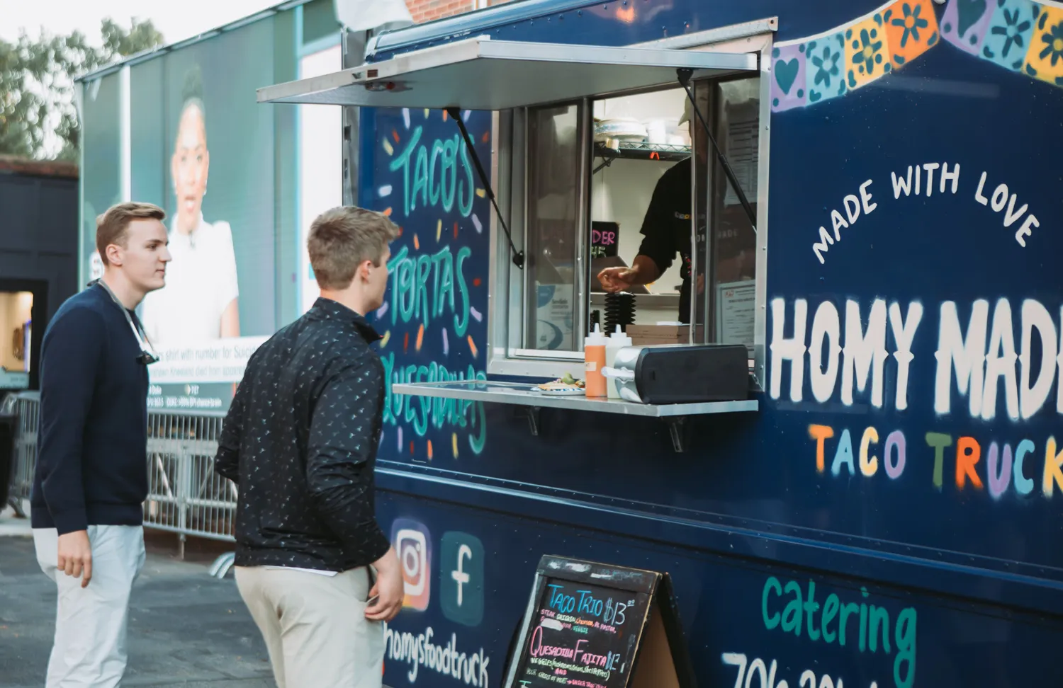 Two men order at a colorful Homy Made taco food truck with menu board and condiments outside.