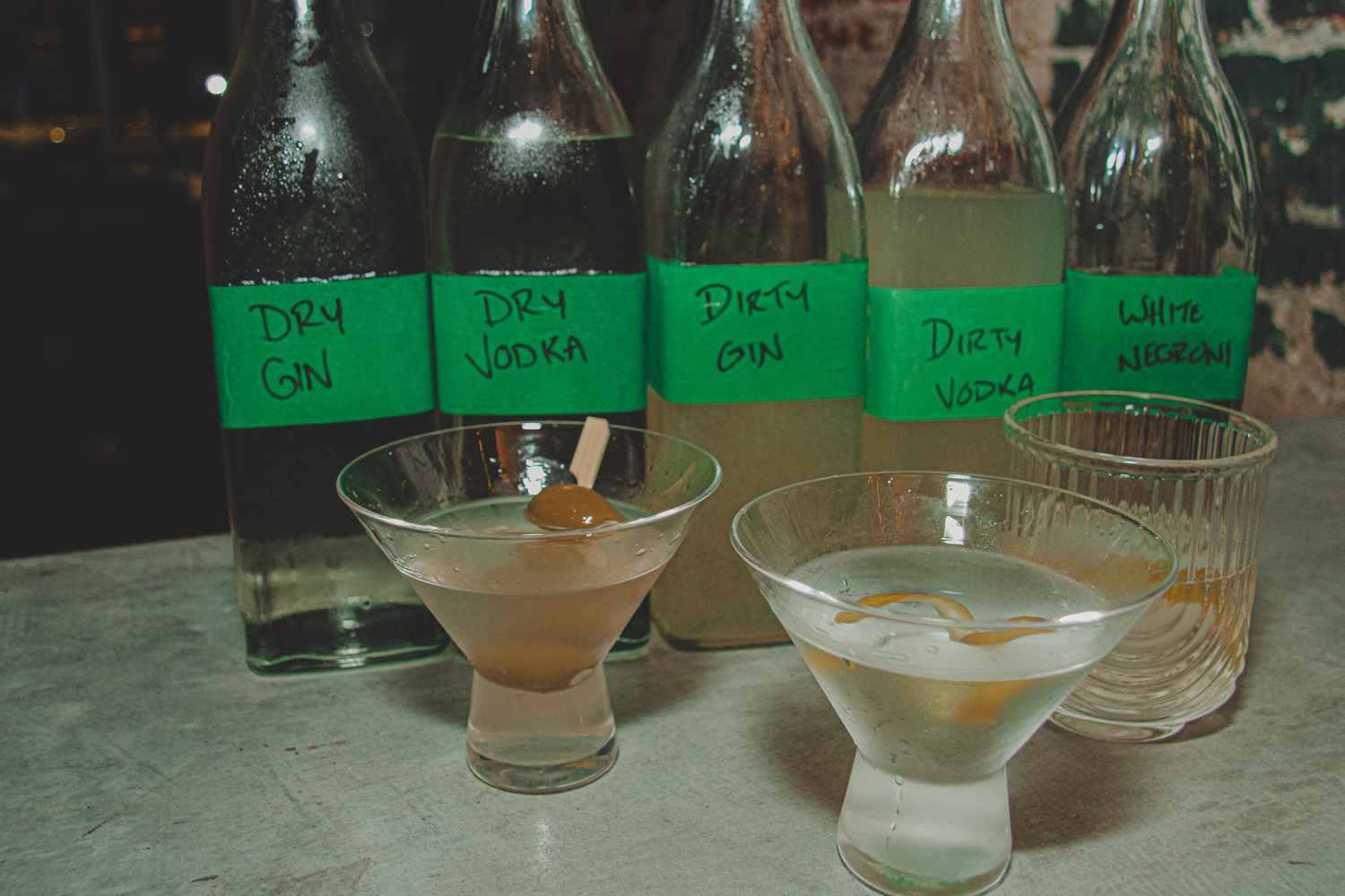 Five bottles labeled with cocktail names and three mixed drinks on a concrete surface.