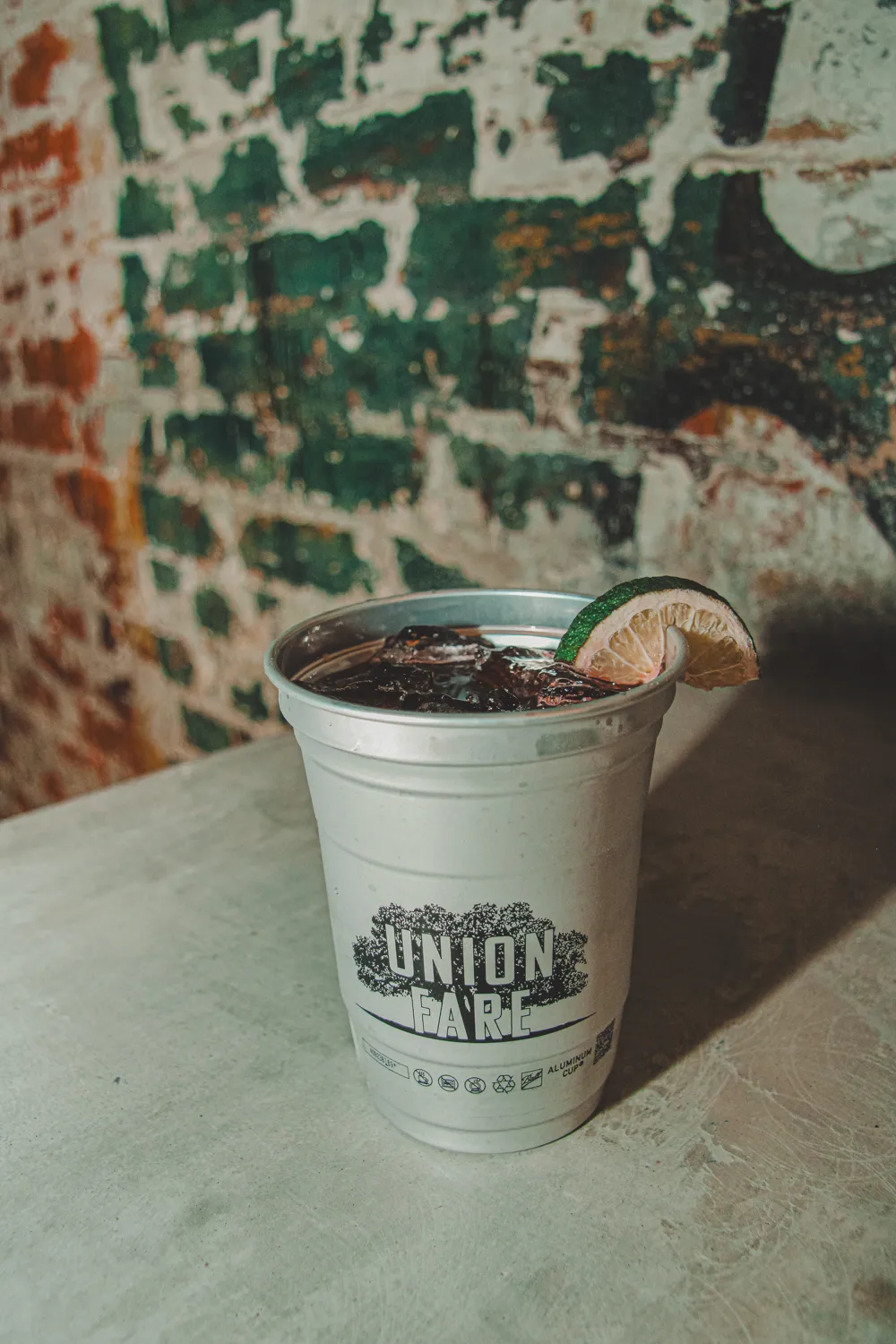 White aluminum cup with 'Union Fare' text holds iced drink garnished with a lime wedge on a table.