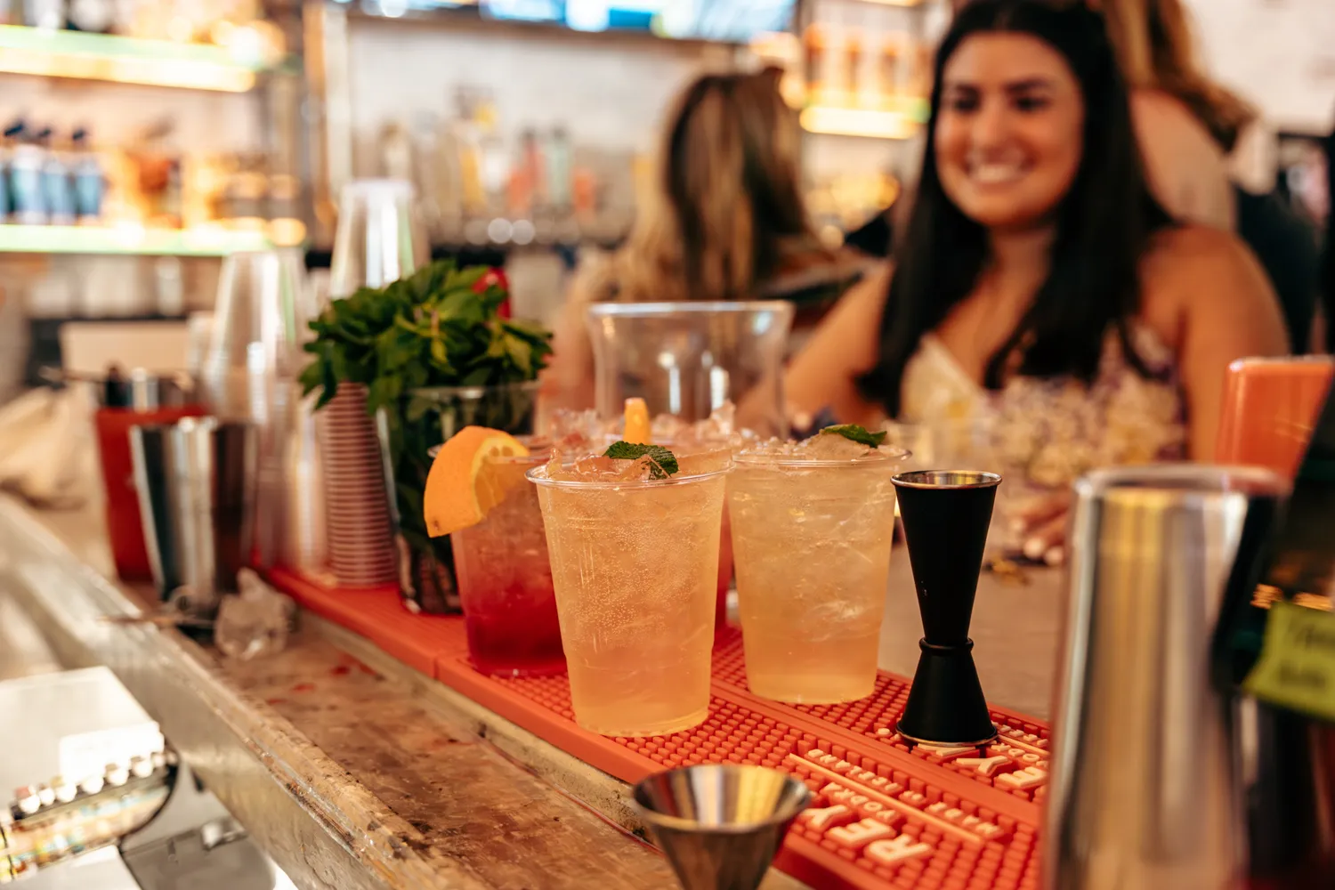 Two iced cocktails garnished with mint and lemon on a bar counter with a smiling woman blurred in background.