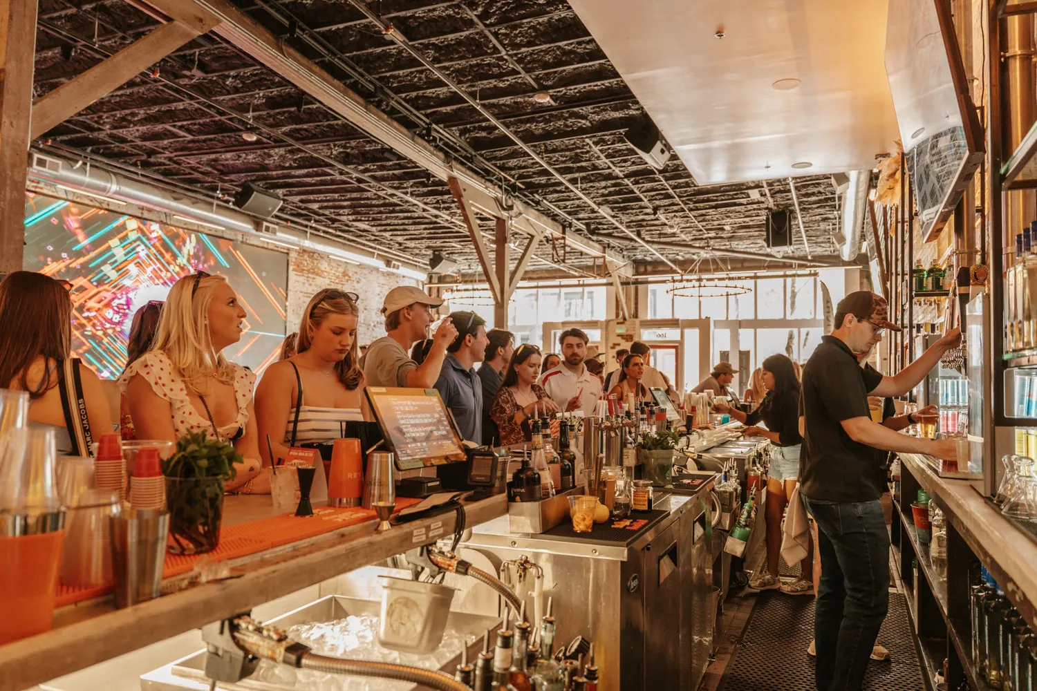 Busy bar with patrons standing and bartenders serving drinks behind counter in a well-lit rustic space.