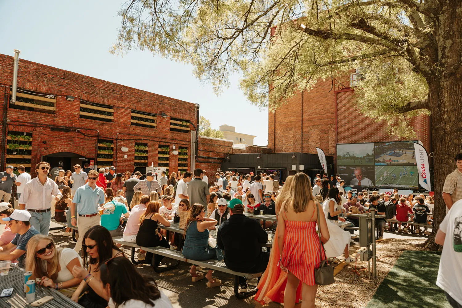 Crowd socializing at outdoor venue with picnic tables, large screen showing sports, and brick buildings.