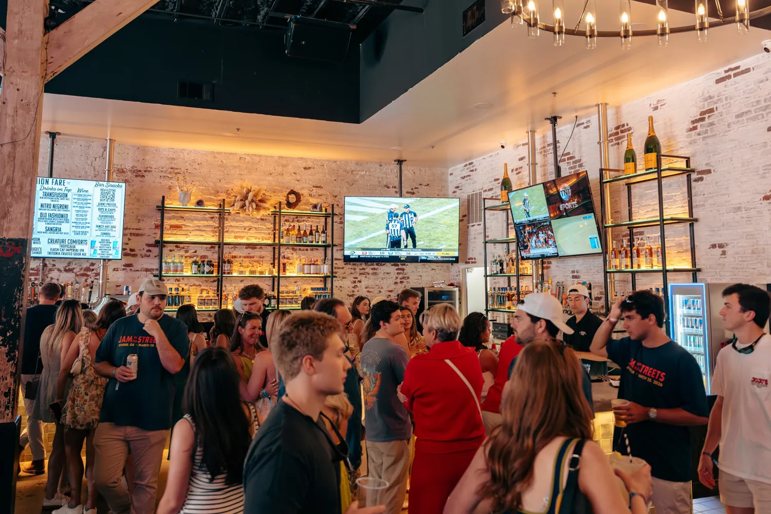 Crowded bar with people socializing, illuminated shelves with bottles, and TVs showing a sports game.