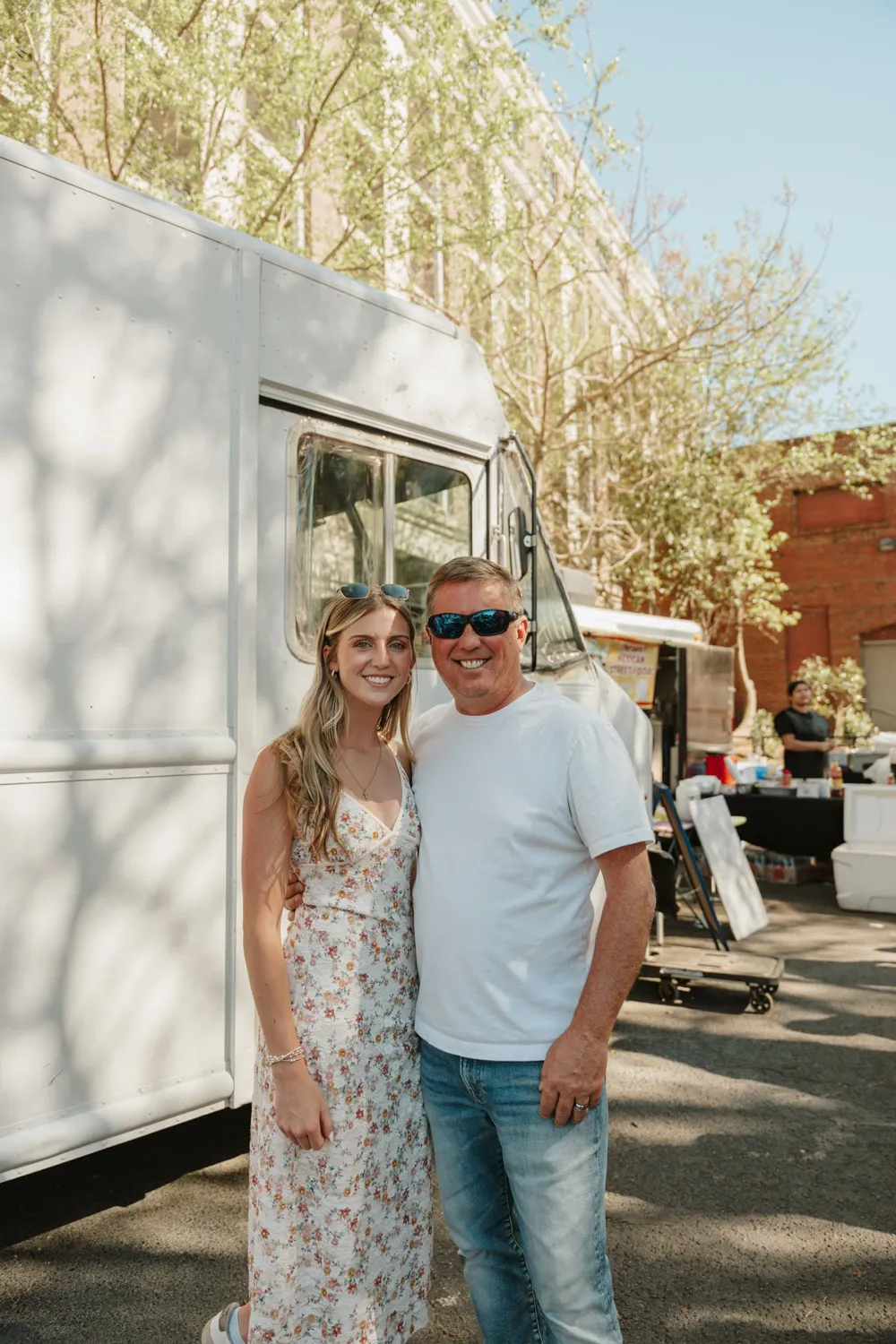 Smiling man in sunglasses and woman in floral dress standing near a white food truck outdoors.