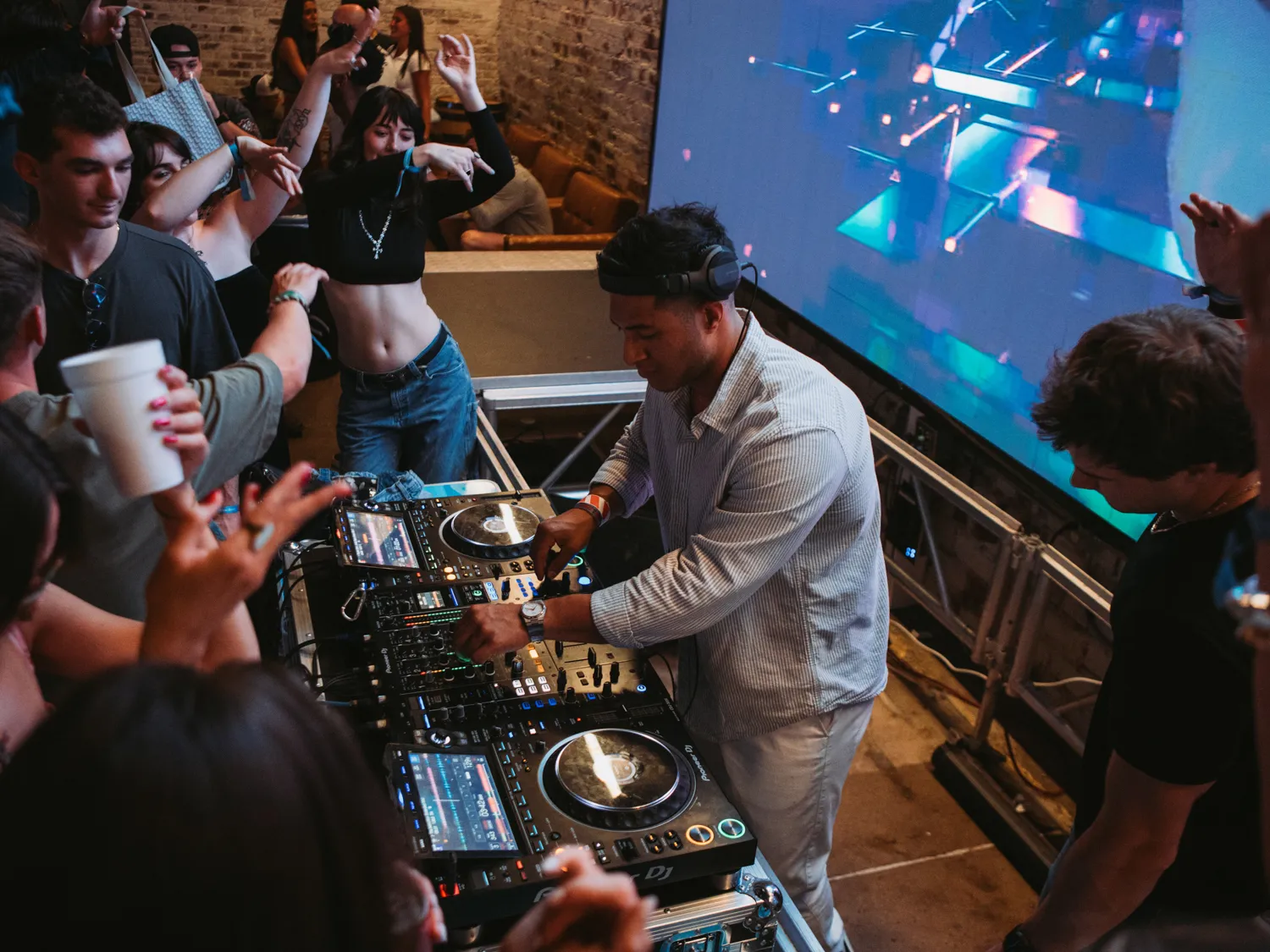 DJ in headphones mixing music at a party as people dance and raise hands nearby.