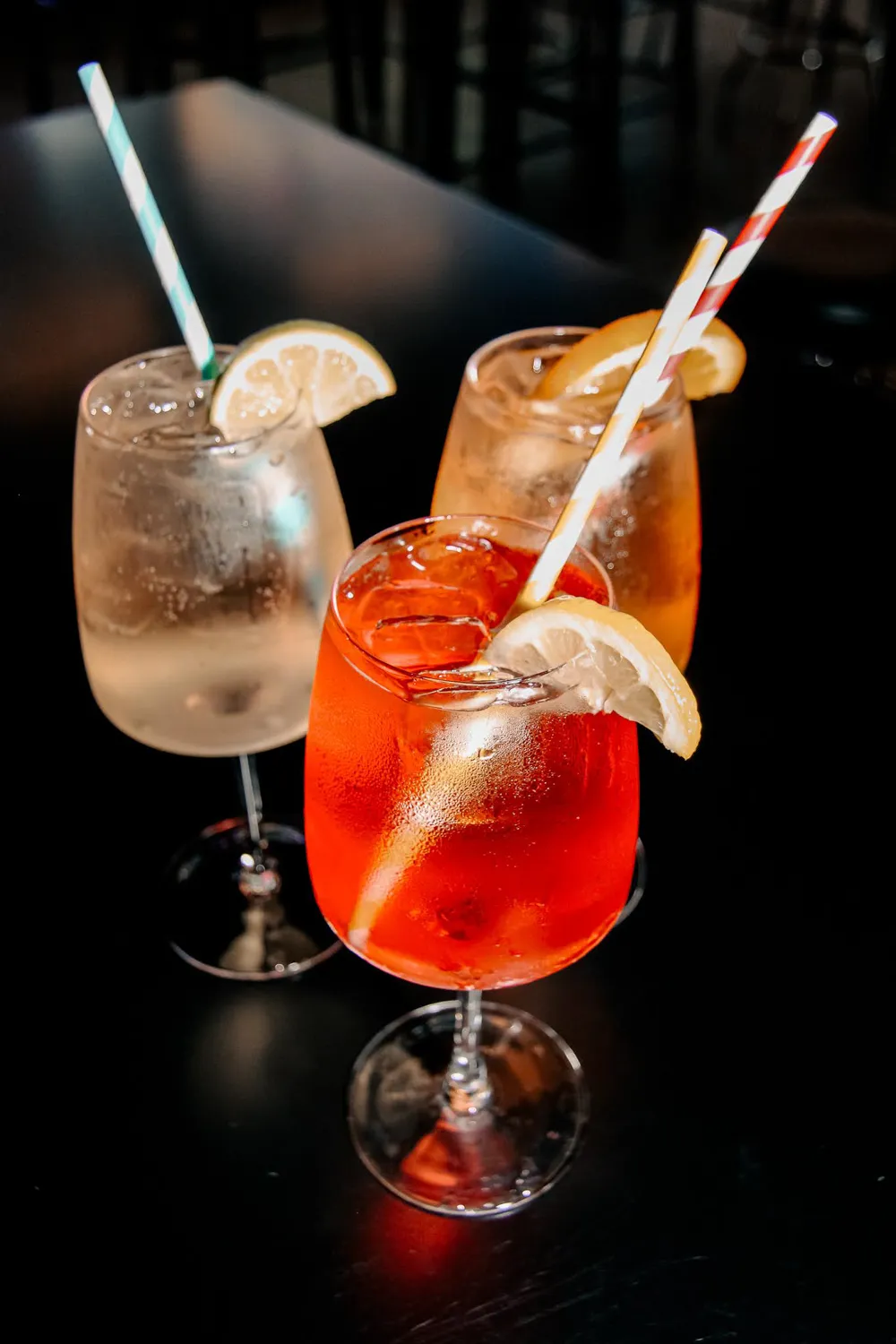 Three iced cocktails garnished with lemon slices and striped straws on a dark table.