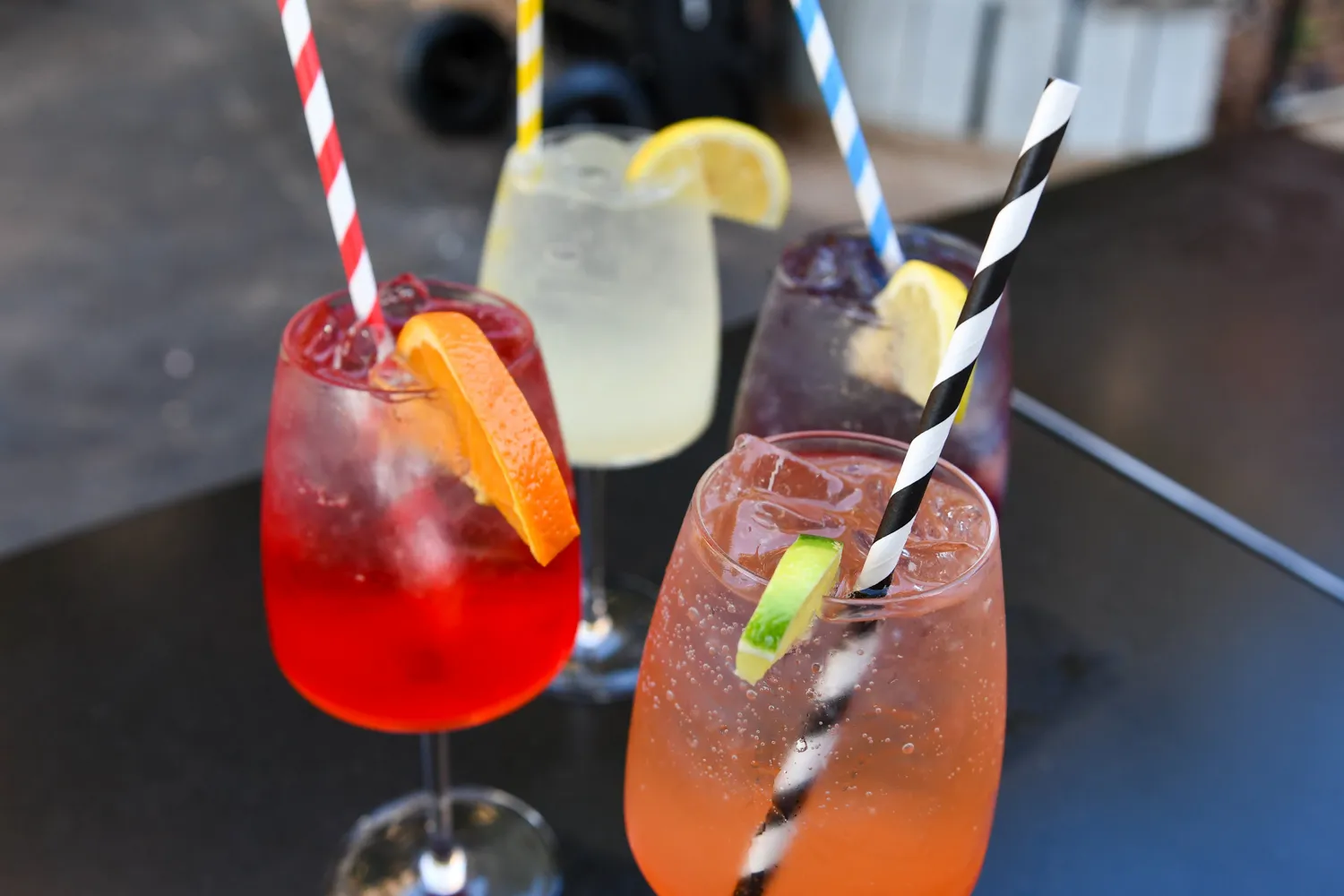 Four colorful cocktails with ice and citrus garnishes on a black table outdoors.