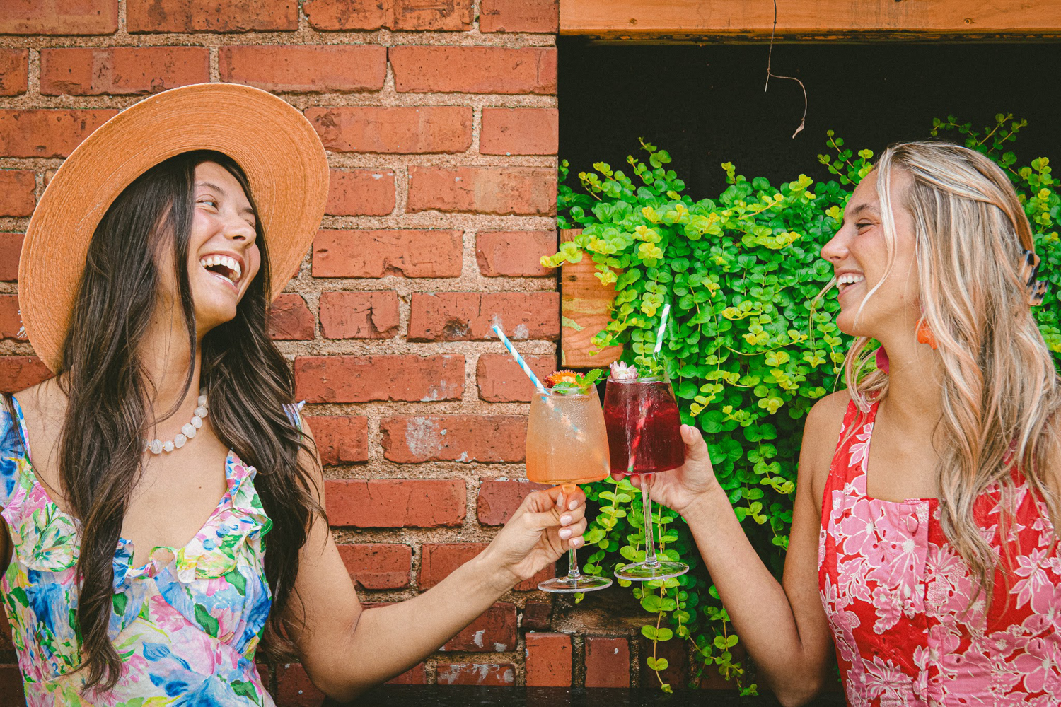 Two women in colorful dresses clinking cocktail glasses together, smiling against a brick wall.