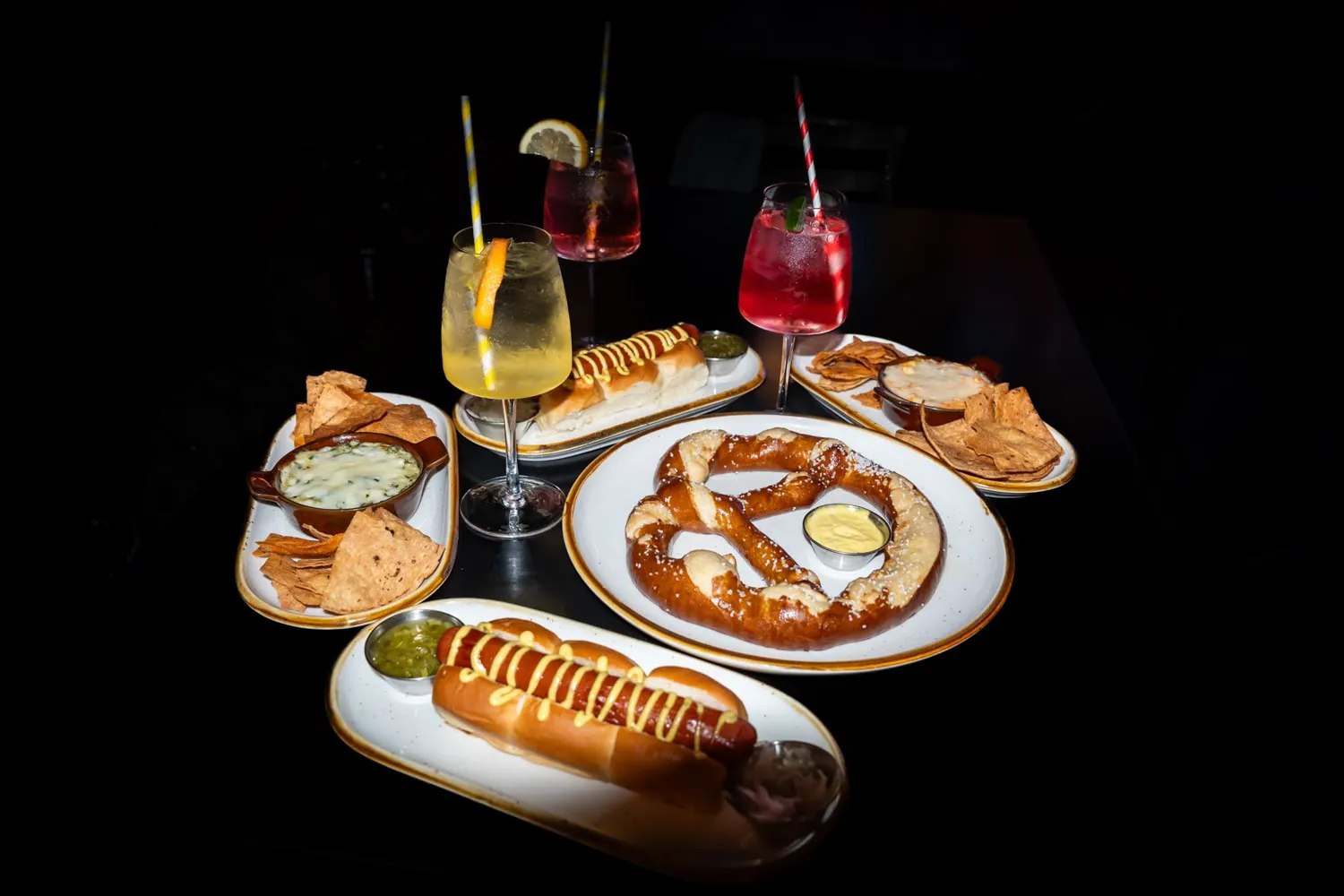 Plates of soft pretzel, hot dogs with mustard, chips with dips, and three colorful cocktails on a dark table.