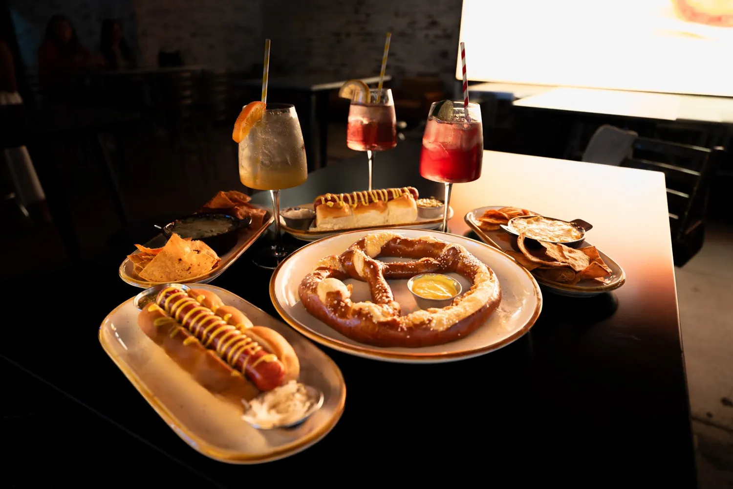 Table with pretzel, hot dogs, chips with dips, and three cocktails with fruit garnishes.