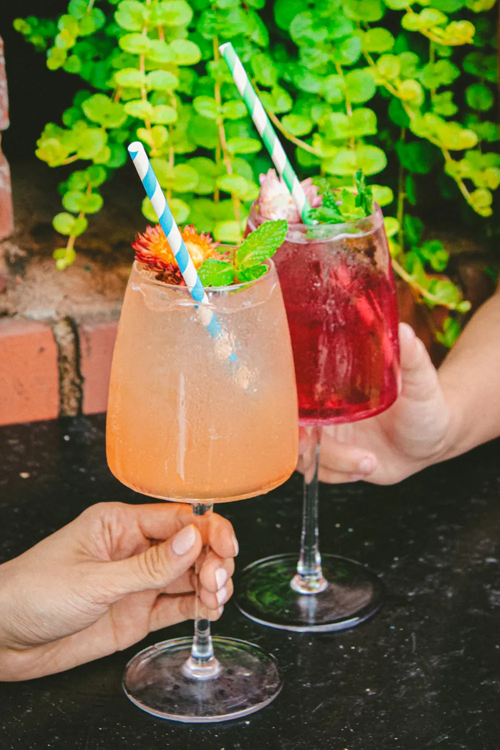 Two hands holding colorful cocktails with striped straws and mint garnish outdoors.