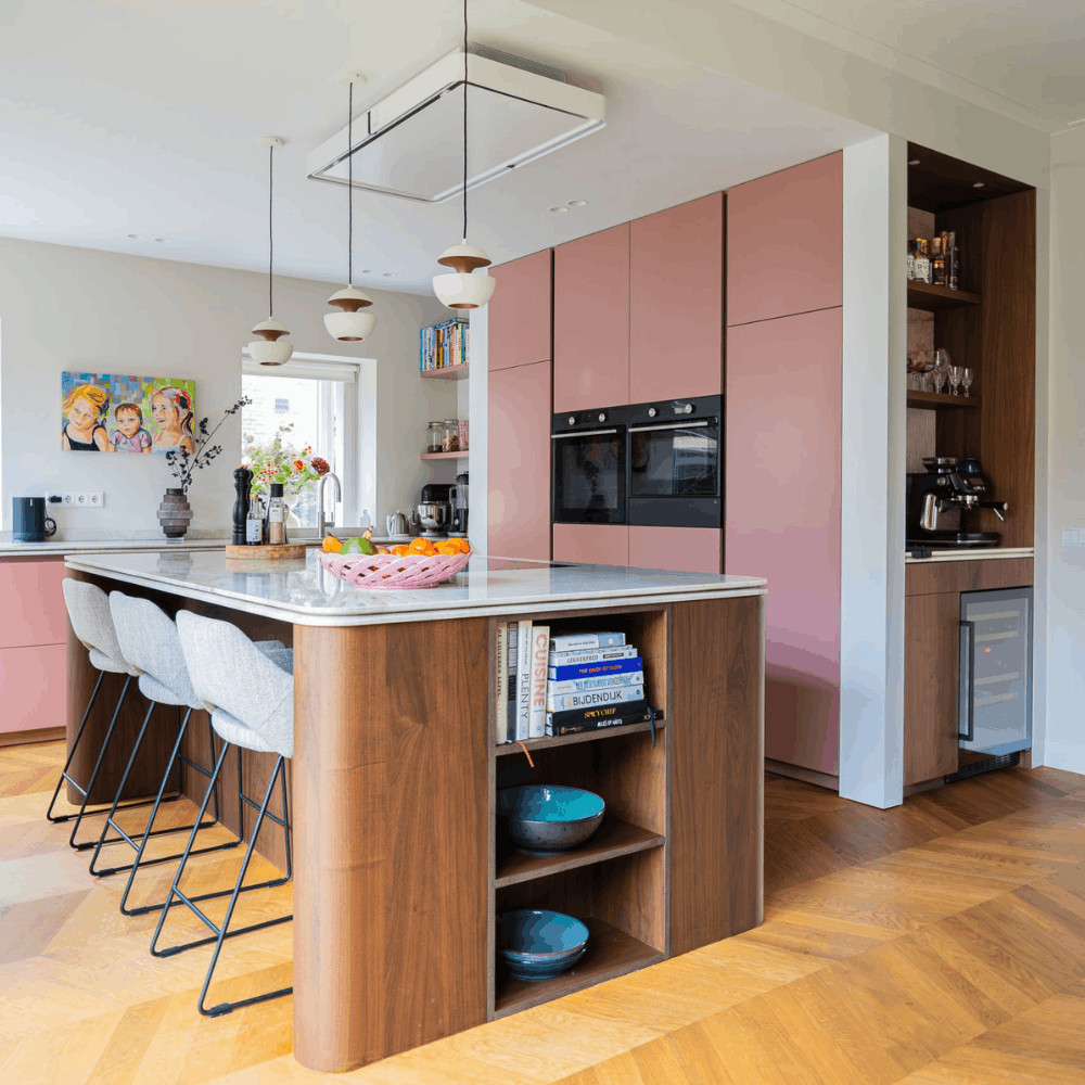 Moderne keuken met roze kastdeuren, marmeren eiland met drie grijze barkrukken, open houten planken met kookboeken en keukenapparatuur.