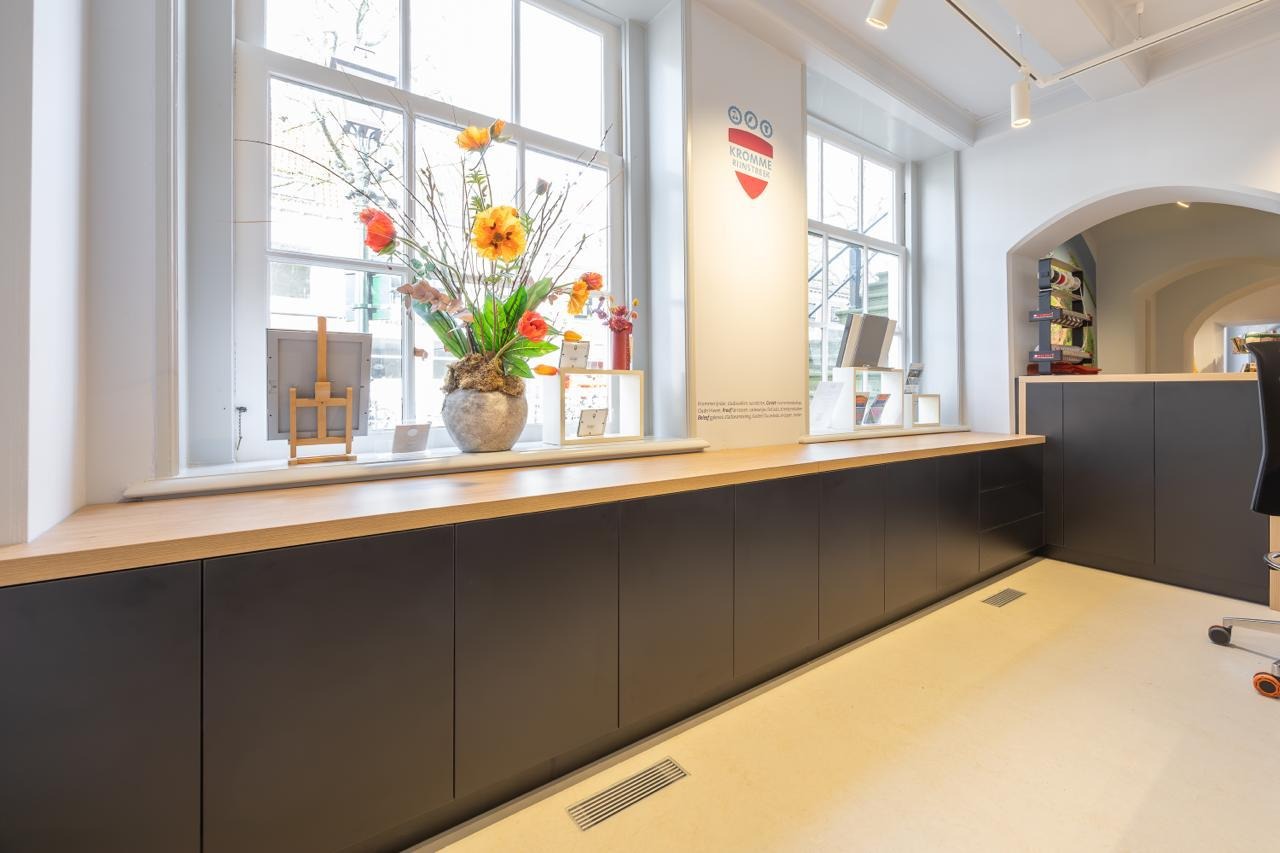 Lange zwarte dressoirkast met houten bovenblad onder grote ramen met bloemstuk en decoraties in een lichte winkelruimte.