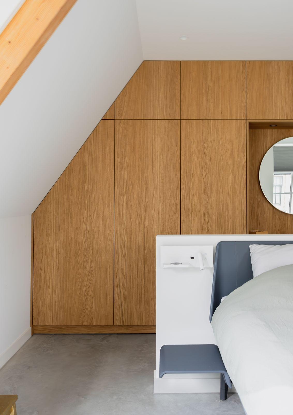 Slaapkamer met schuine wand en ingebouwde houten kast naast een wit bed met een grijs nachtplankje.