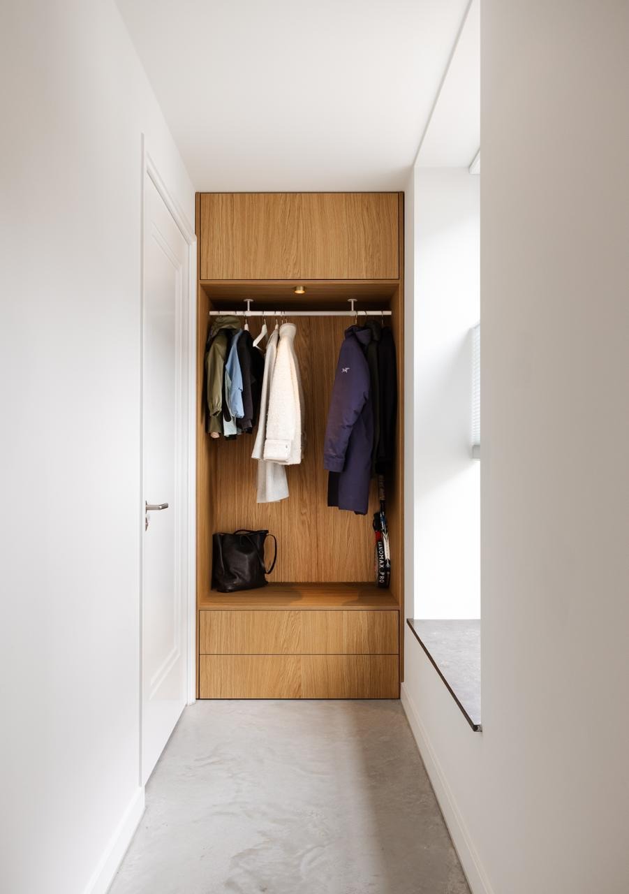 Minimalistische gangkast van licht hout met jasjes, een zwarte tas en een paraplu, tegen een witte muur.