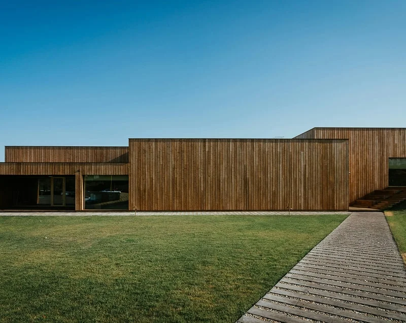 Modern wooden house with horizontal slats, large windows, and a wooden pathway leading through a green lawn under a clear blue sky.