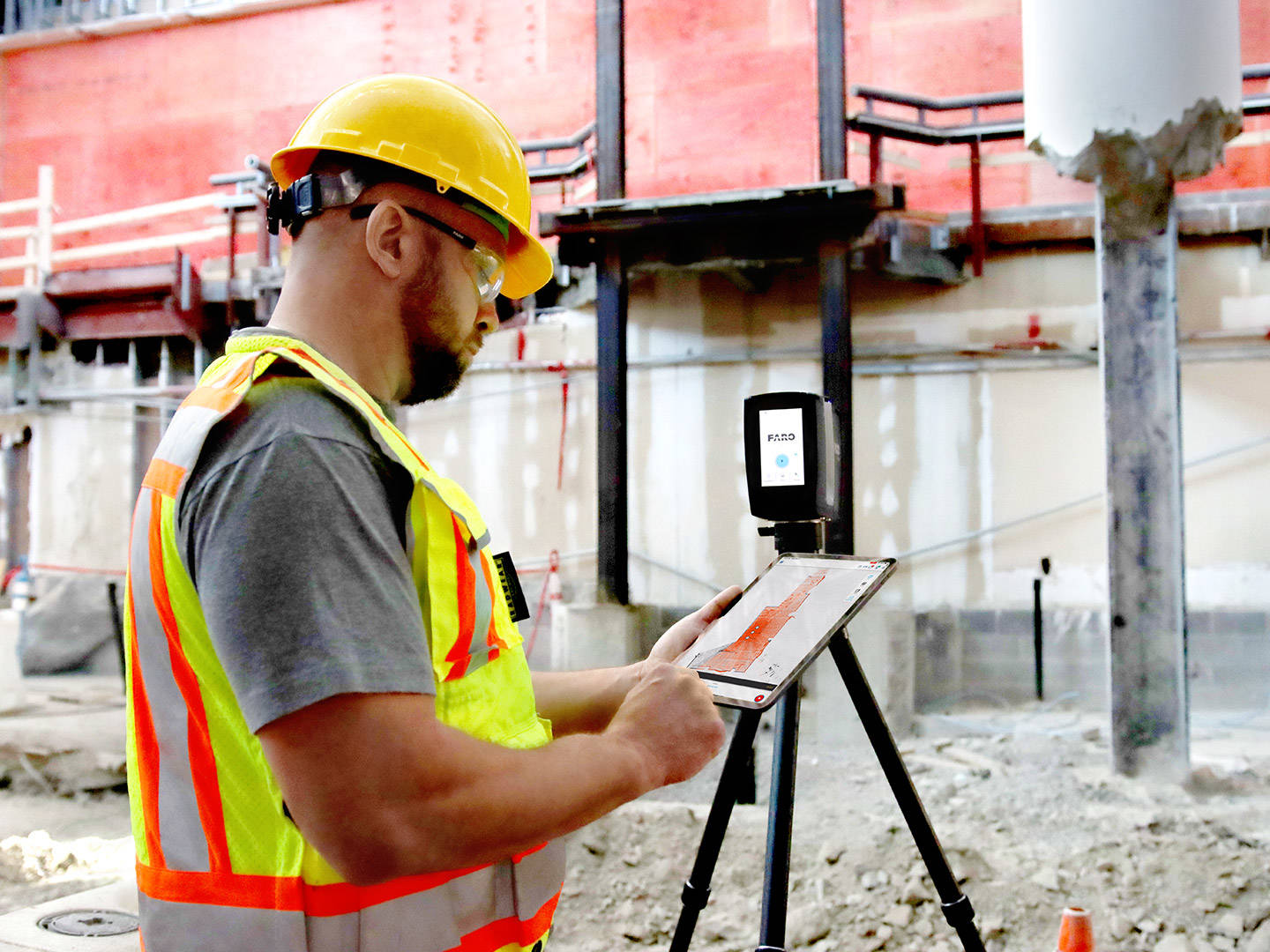 Construction worker in safety vest and helmet using a tablet mounted on a tripod in an indoor construction site.