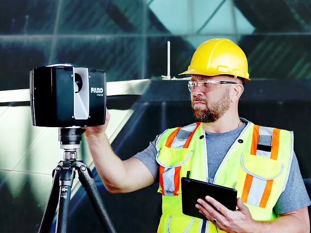 Construction worker in a yellow hard hat and high-visibility vest using a FARO Focus 3D scanner on a tripod while holding a tablet.