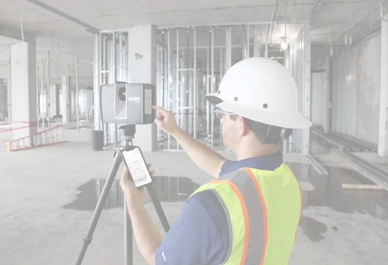 Construction worker in a white hard hat and safety vest using a laser scanner and smartphone inside an unfinished building.