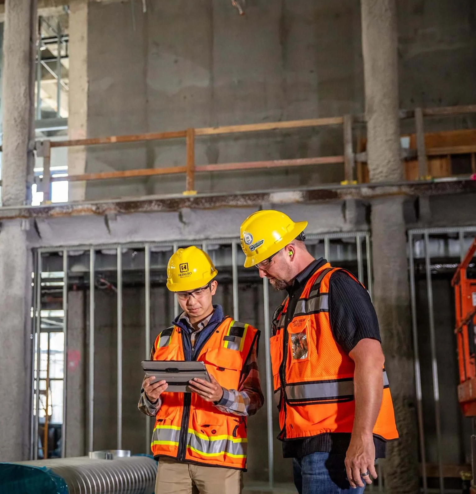 Two construction workers wearing yellow helmets and orange safety vests reviewing a tablet inside a building under construction.