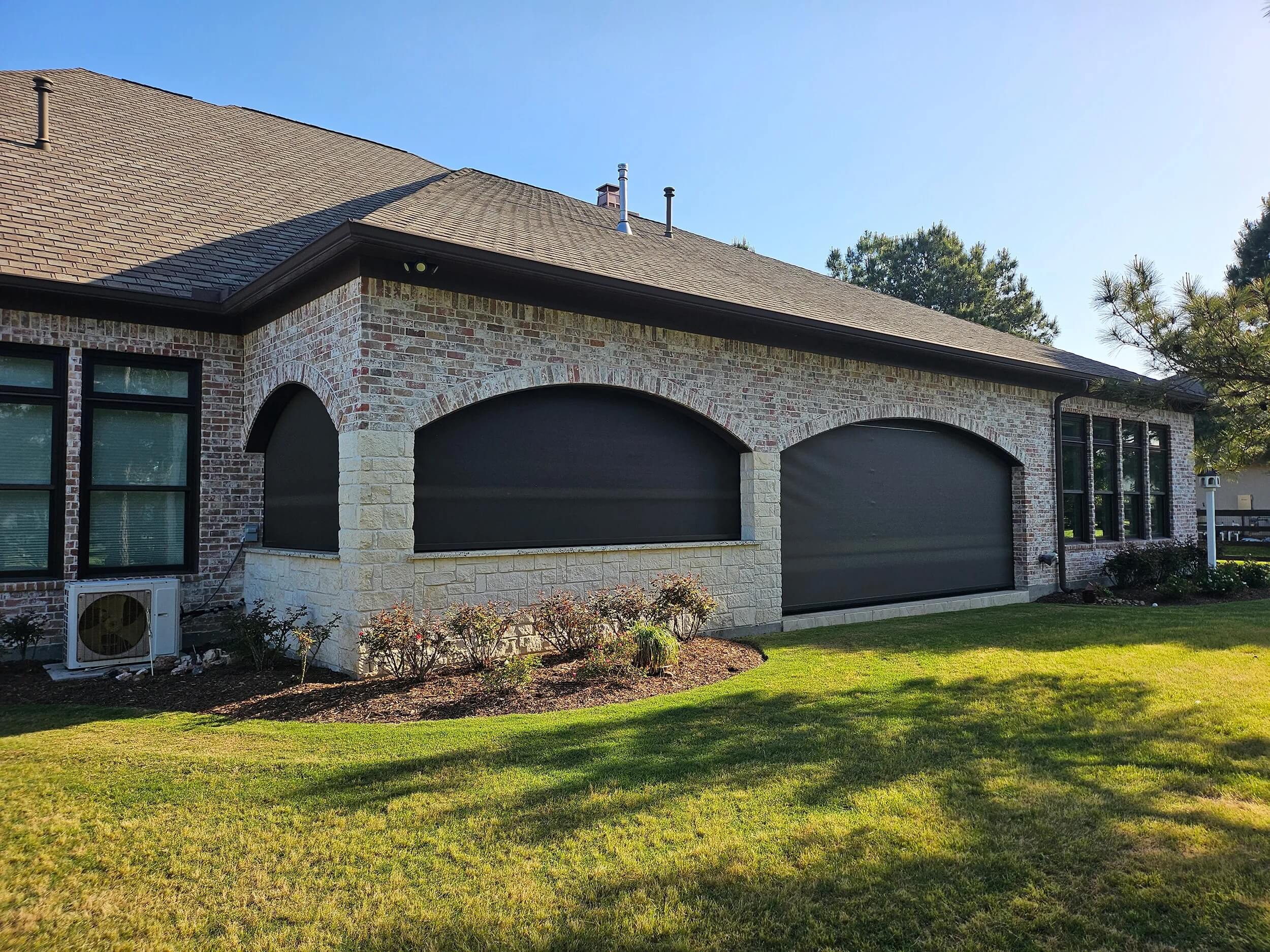 Brick house with beige stone accents, three large arched windows covered by black shades, and a green lawn.